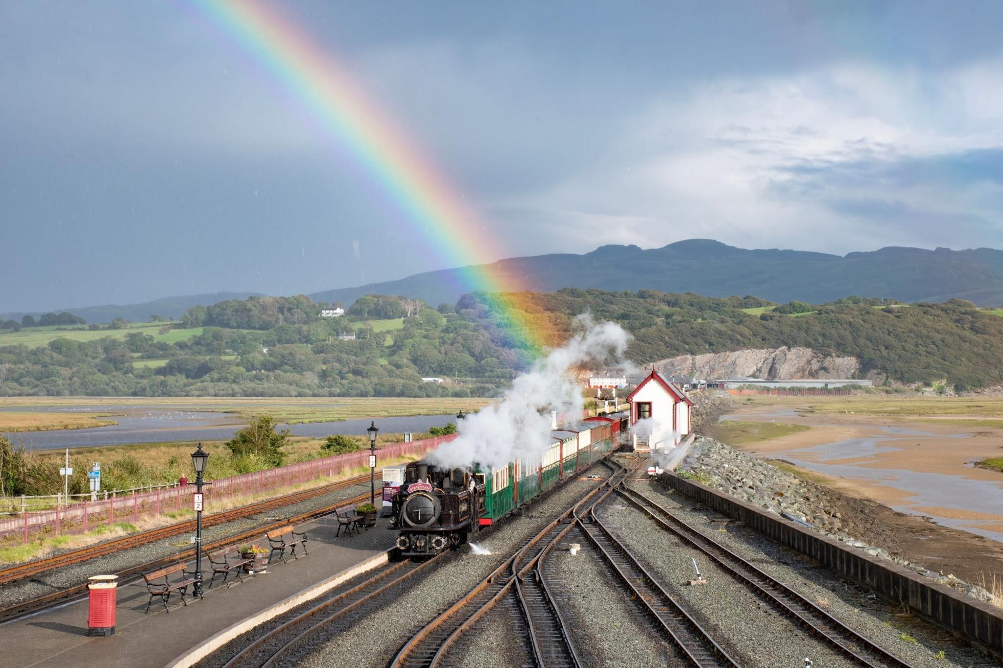 A steam train arrives at a station under a rainbow