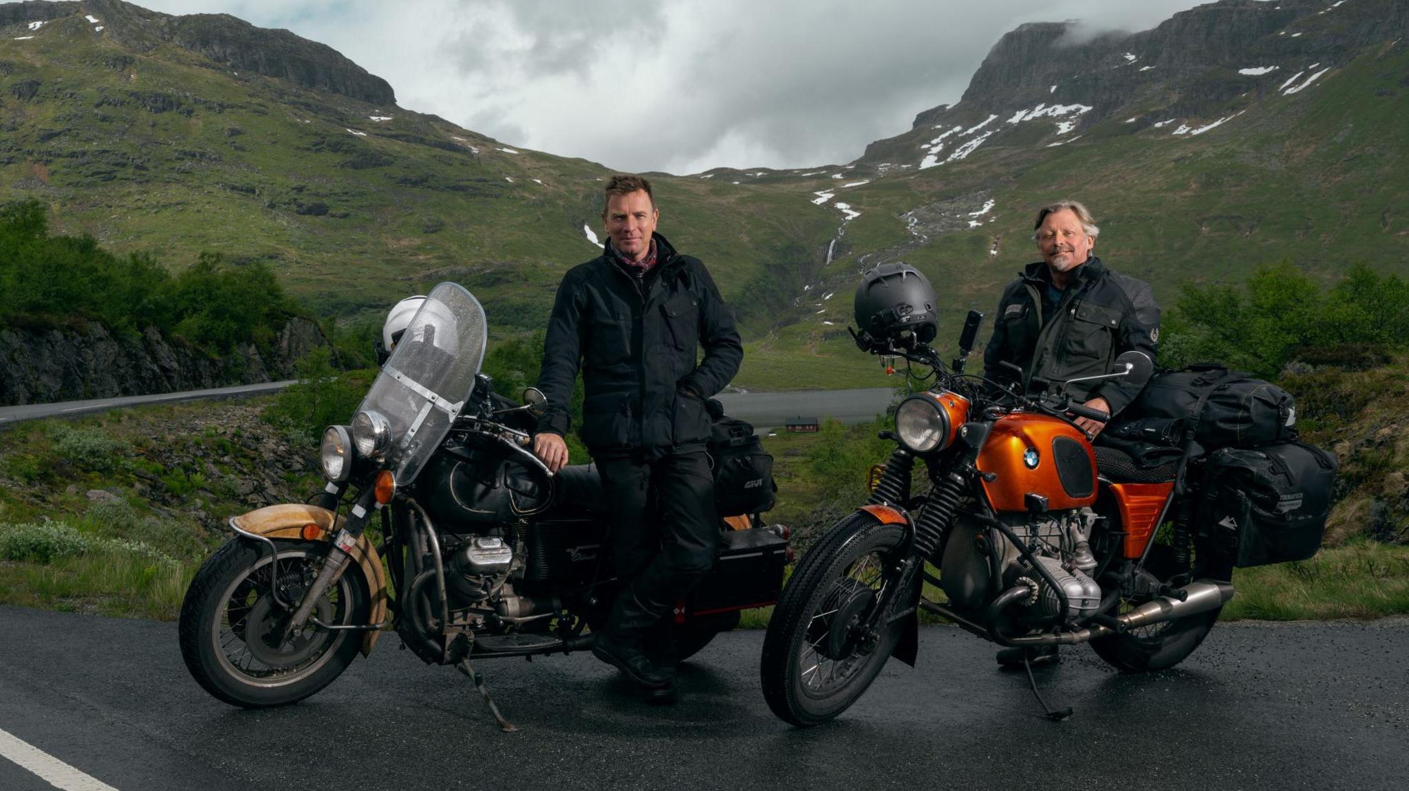 Ewan McGregor and Charley Boorman, leaning on their motorbikes with a stunning snow-peaked glen behind them. Both are in dark biker clothes.