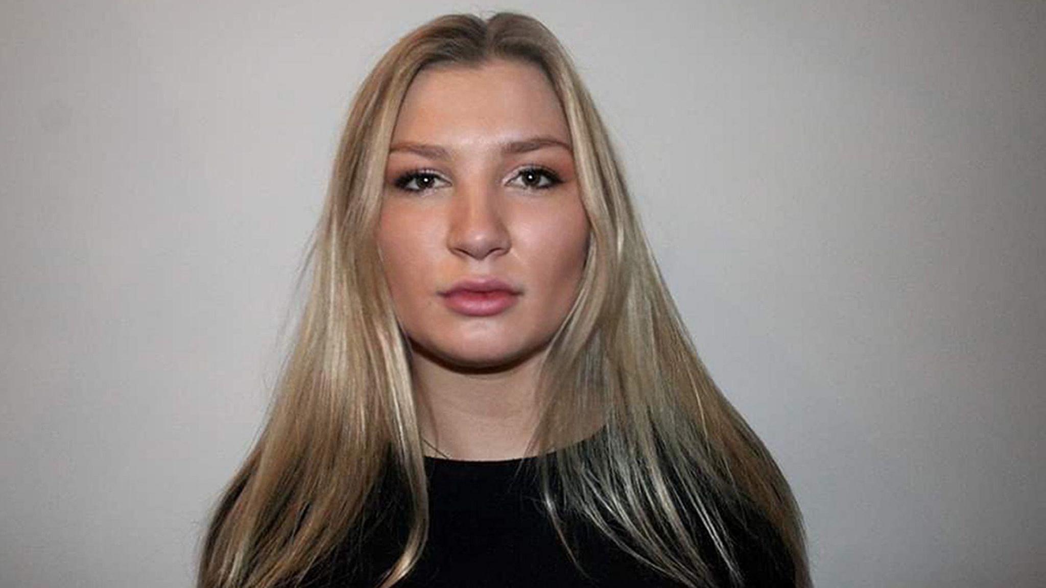 A woman with long blonde hair pictured against a beige wall. She is wearing a black top and is looking at the camera.