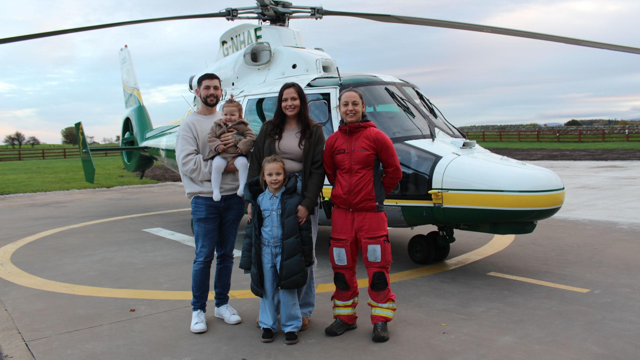 Olivia Brummitt and Dan Atkinson, their daughters and an air ambulance crew member stand in front of Great North Air Ambulance helicopter on a helipad marking.