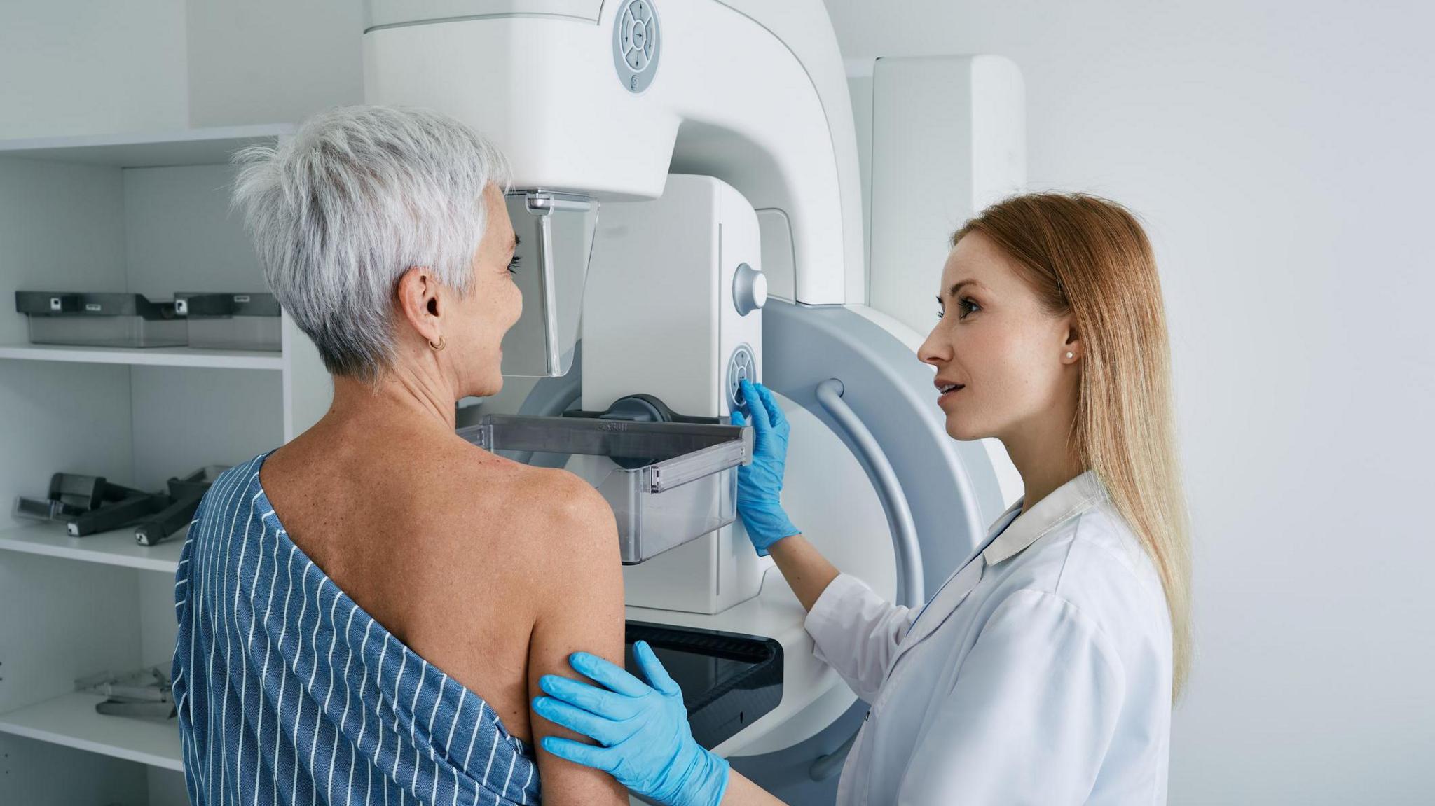 A female worker in a white lab coat and blue gloves screens a lady with short grey hair. The worker has her left arm touching the right arm of the patient. They are looking at each other. The patient has a hospital gown covering half of her upper body. She is standing at the white machine to the front of her. There are white shelves to the left