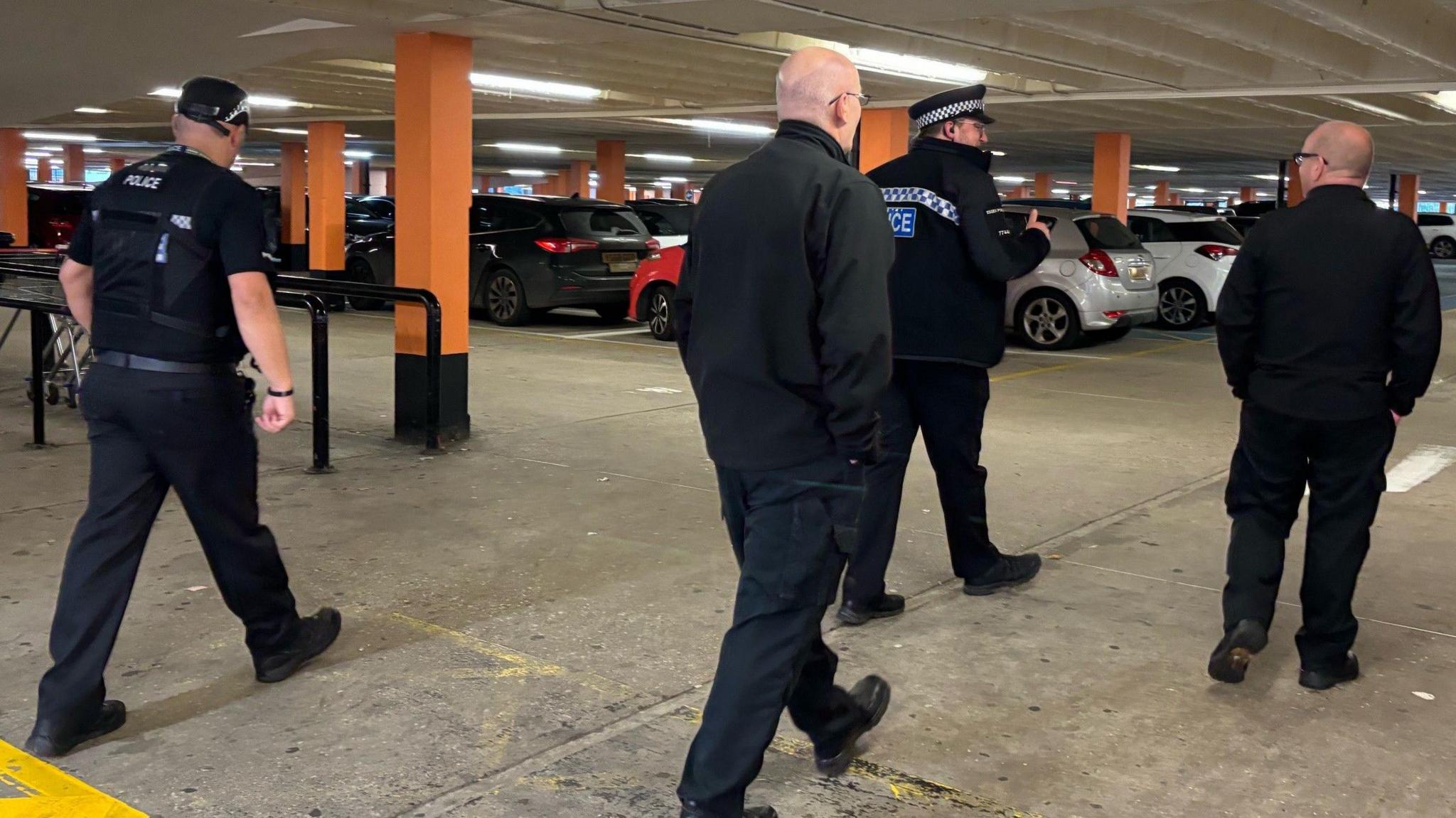 Two Essex Police officers and two men wearing all black walking with their backs turned in a multi-storey car park. There are cars in the background.