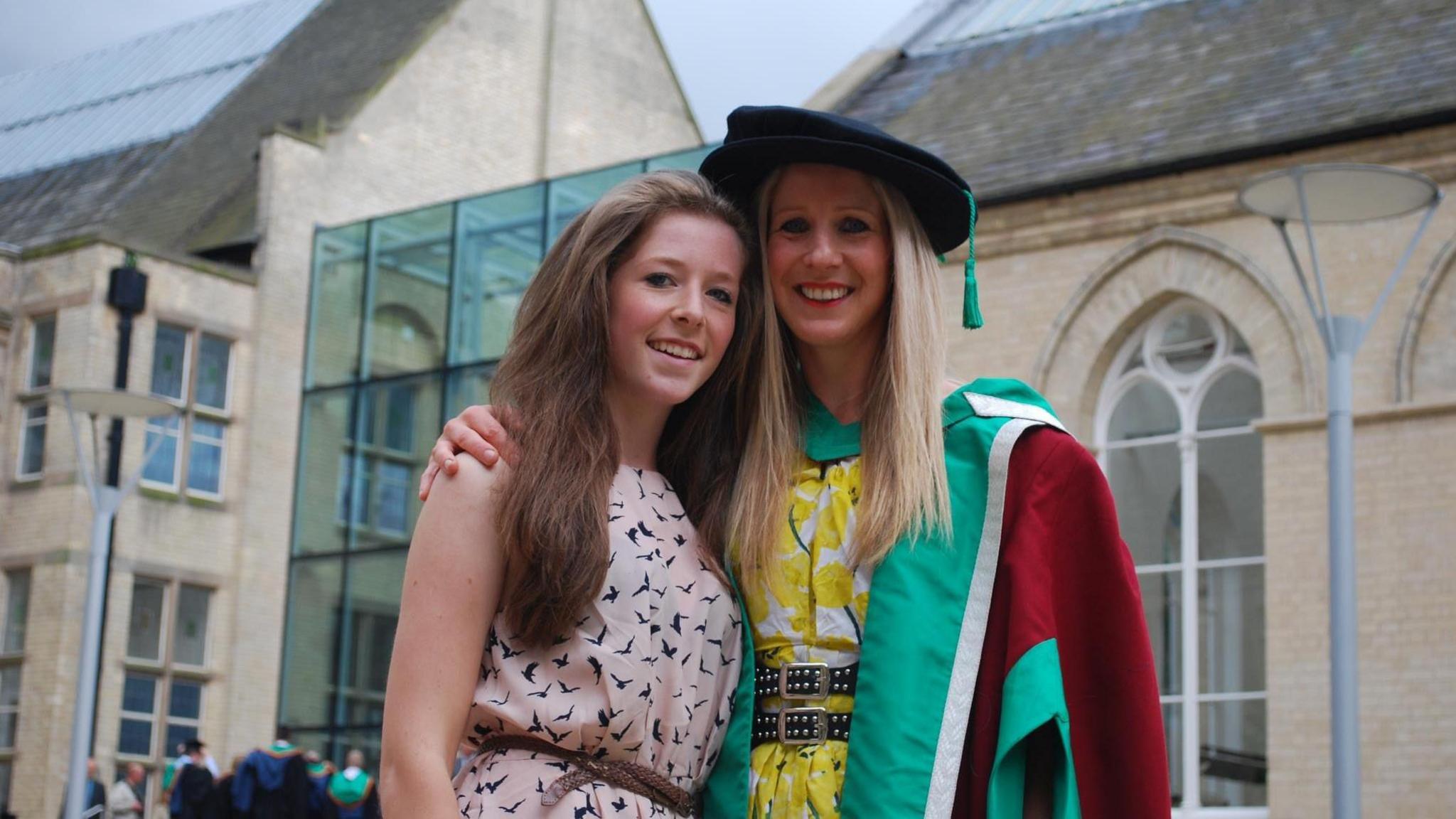 Anna smiles at the camera, wearing a green and red graduation gown and a yellow dress. She has her arm round her daughter Star, who is wearing a patterned light pink dress.