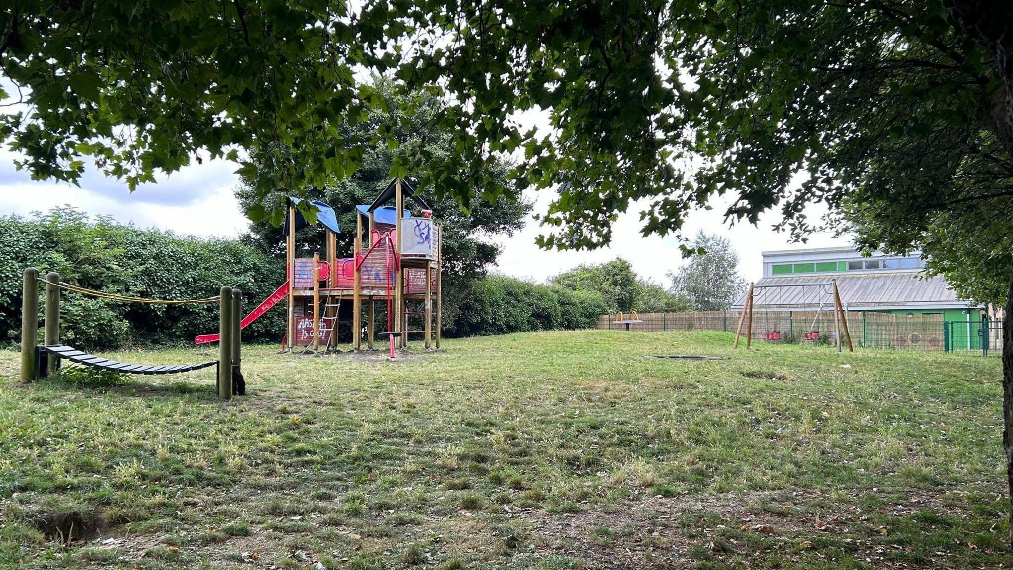 A play area with graffiti on a climbing frame, next to a set of swings and a short wooden bridge