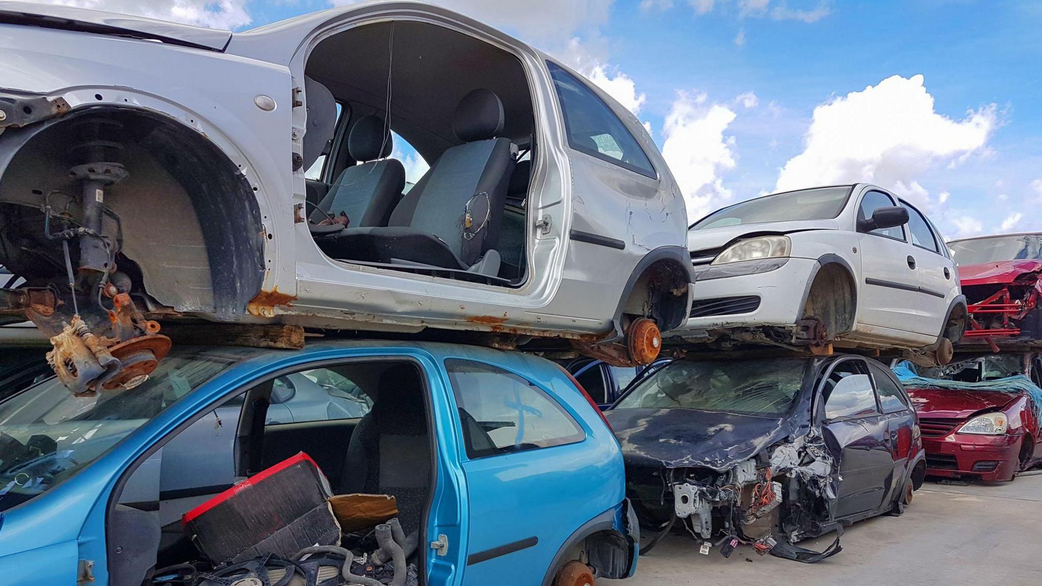 Scrapped cars are piled on top of each other in a row. The wheels are missing and the old cars are rusting and missing components