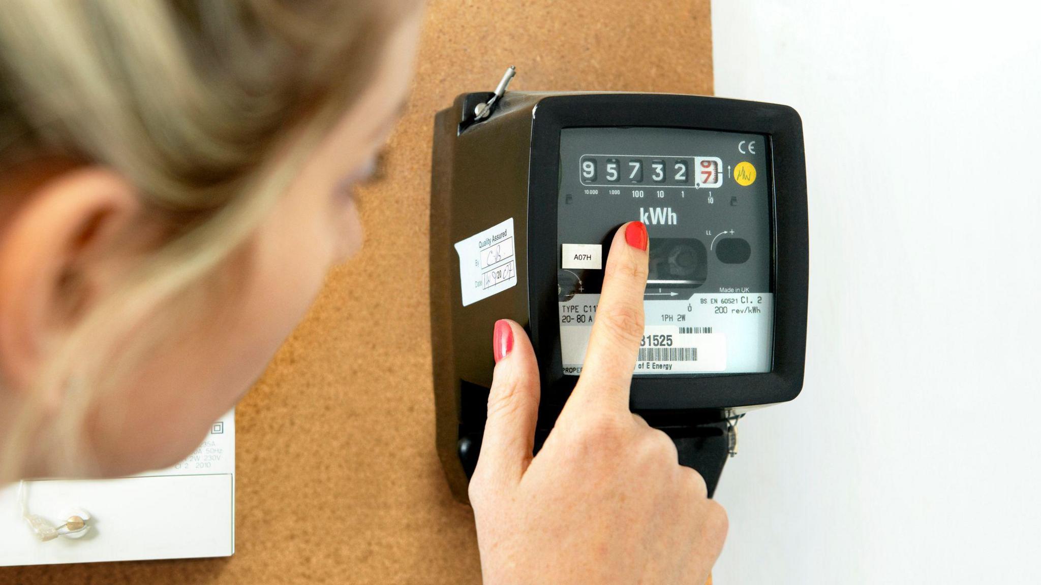 A blonde woman looks at her black electricity metre which is placed on a white wall with a cork board on it. 