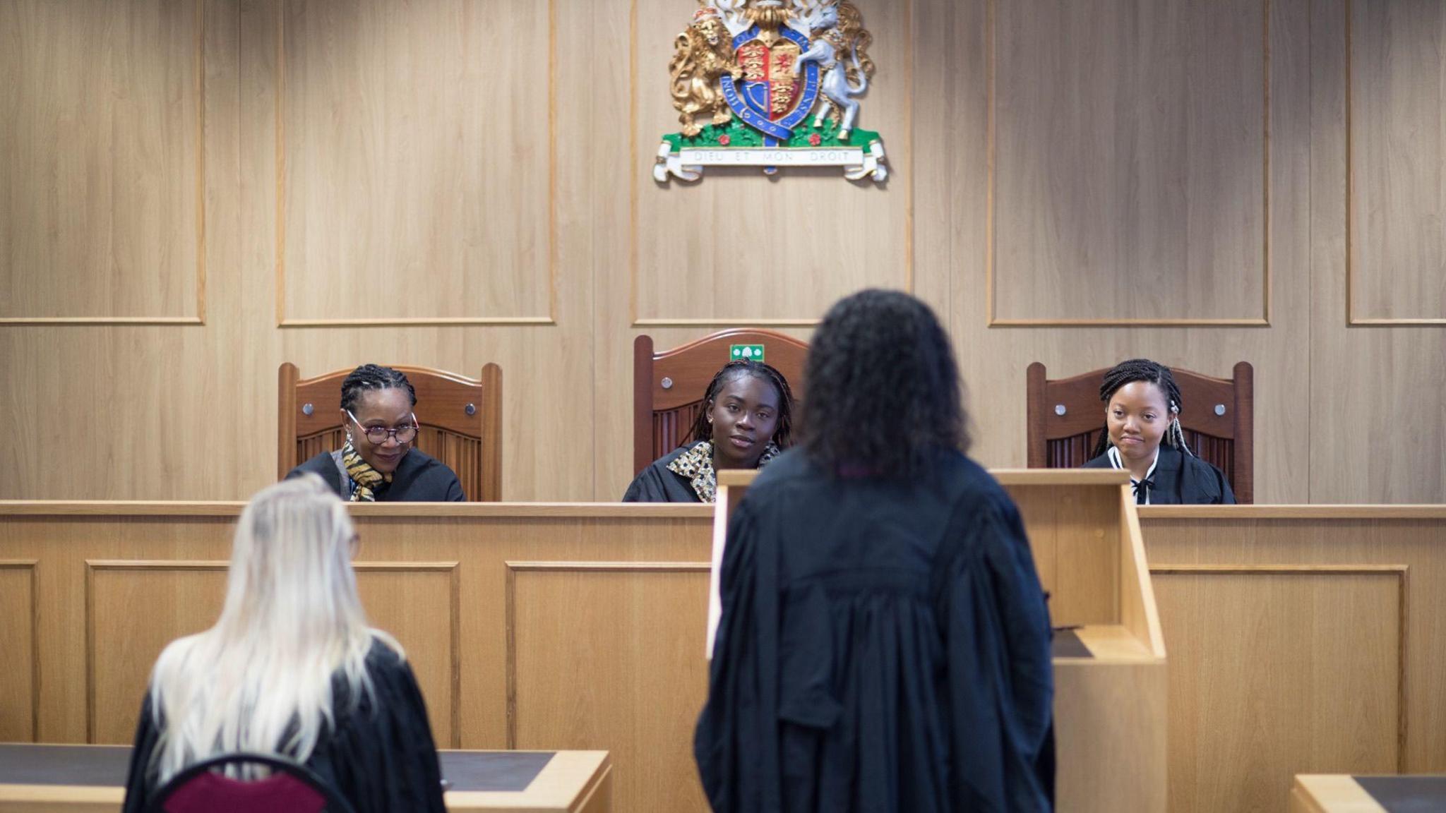 The picture shows students in a wood panelled court room. Three women are sitting in large wooden chairs behind a large desk and facing the camera. There is a crest on the wall above them. Two people are facing them, with their backs to the camera wearing black robes. One is sitting at a lower desk, the other is standing at a podium.
