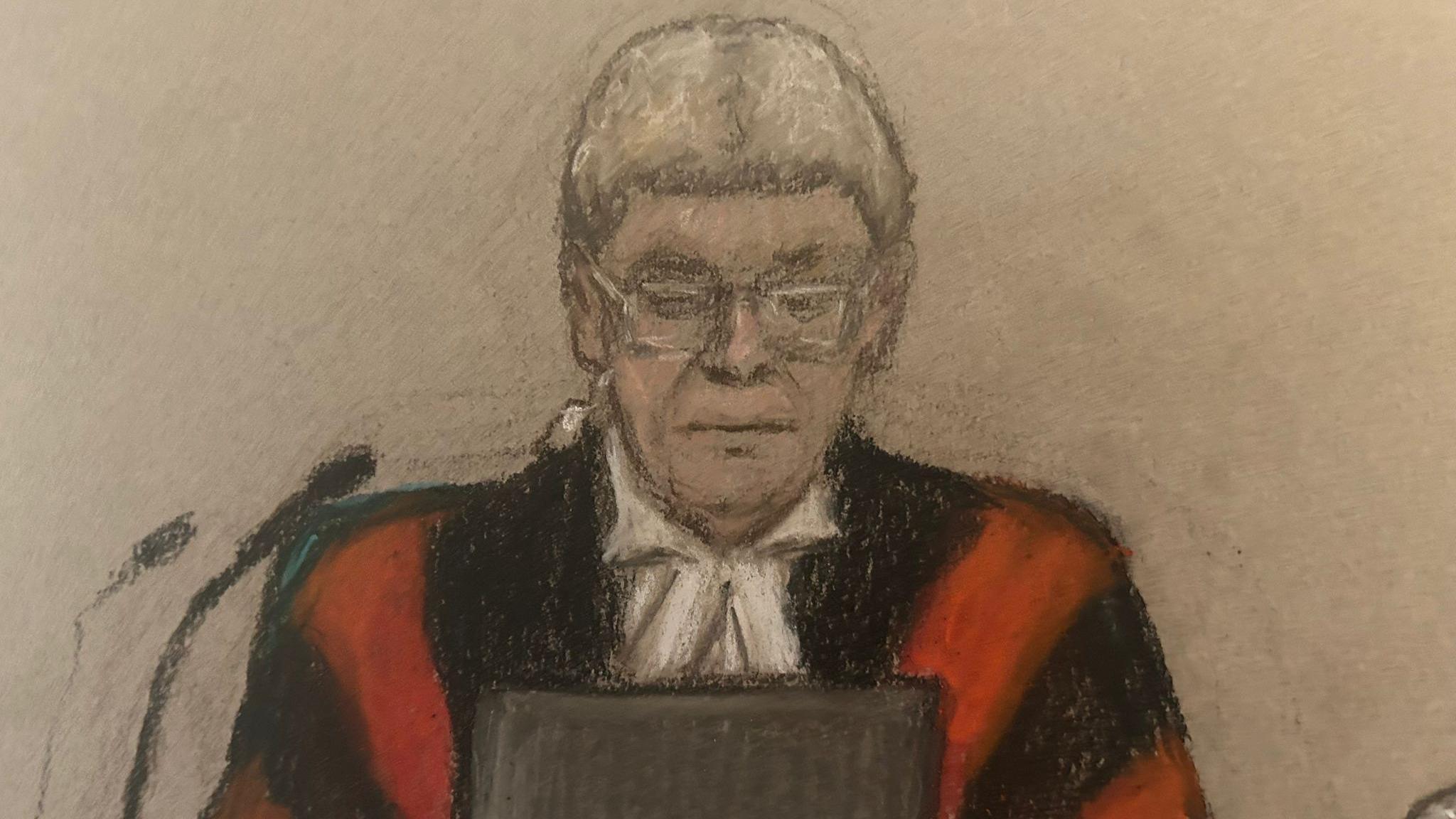 Judge Mark Lucraft, who is wearing red and black judges robes, a white legal wig and glasses. He is looking at a tablet in front of him and there are two microphones to the side