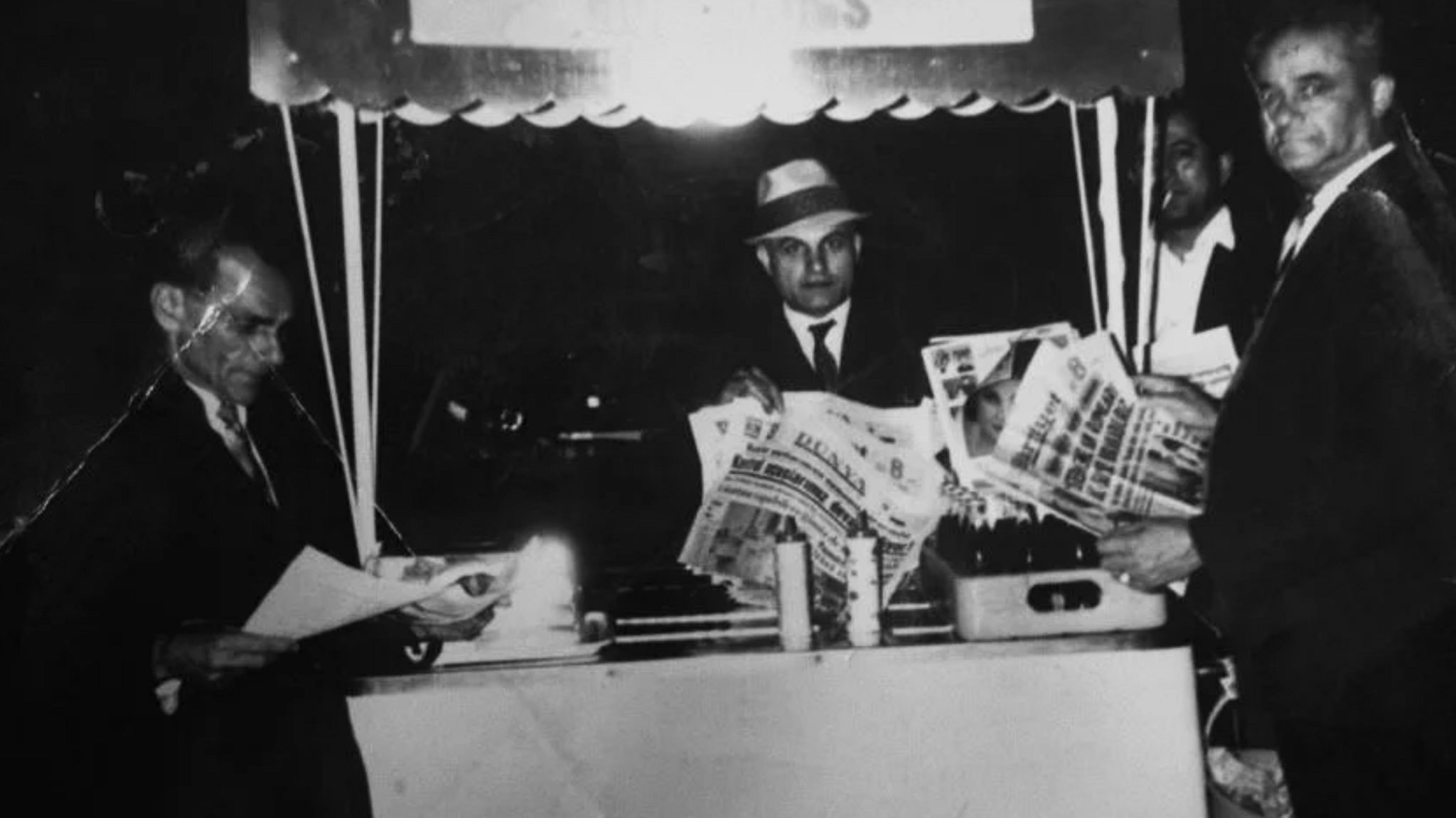 A black-and-white photograph of a street stall at night. Several people stand around the counter, with one person behind the stall holding newspapers. Bottles and other items are displayed on the counter beneath a striped canopy.