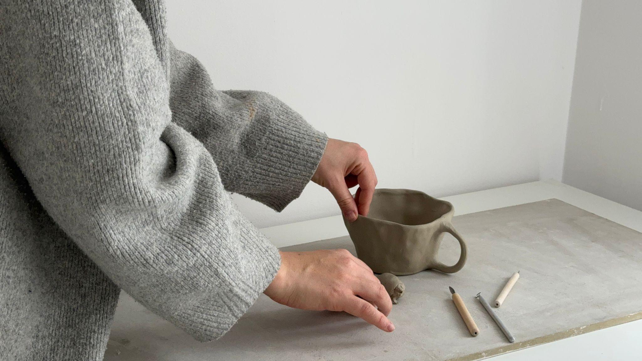 An image of a woman's hands working on an unfired piece of pottery in the shape of a cup, alongside some tools on a bench