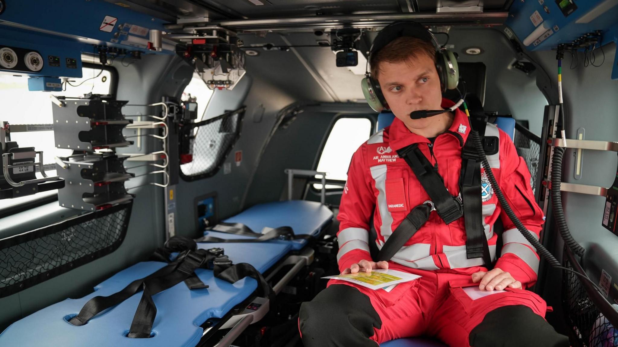 Matthew Keenan, a young man with short hair, is sitting in the new helicopter with a stretcher and equipment next to him. He is wearing a red paramedic uniform and headphones.