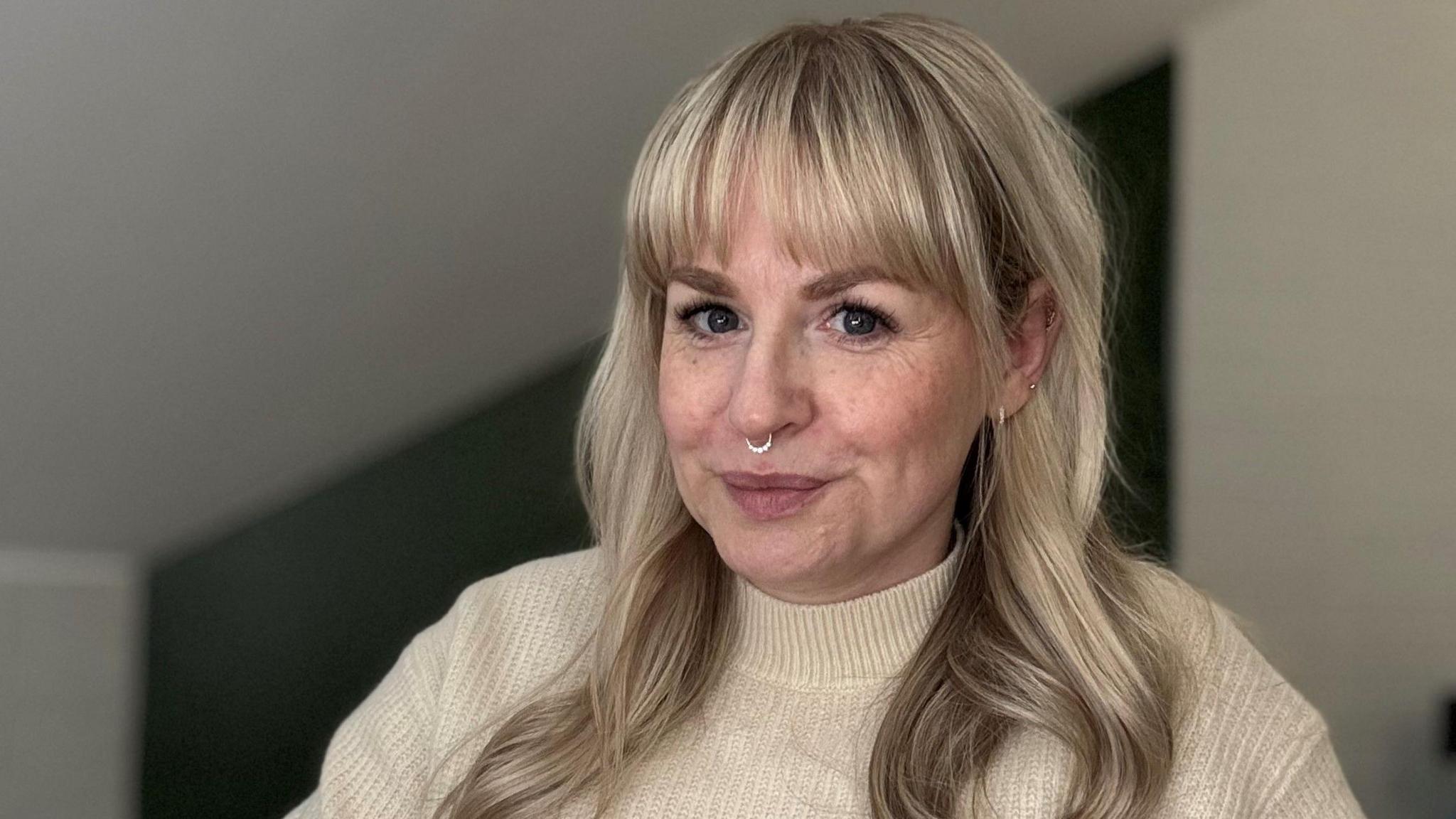 Katie is sitting at her desk. She is wearing a cream turtleneck jumper and has a nose ring. Her hair is long and a silvery, blonde colour, with a straight fringe. 