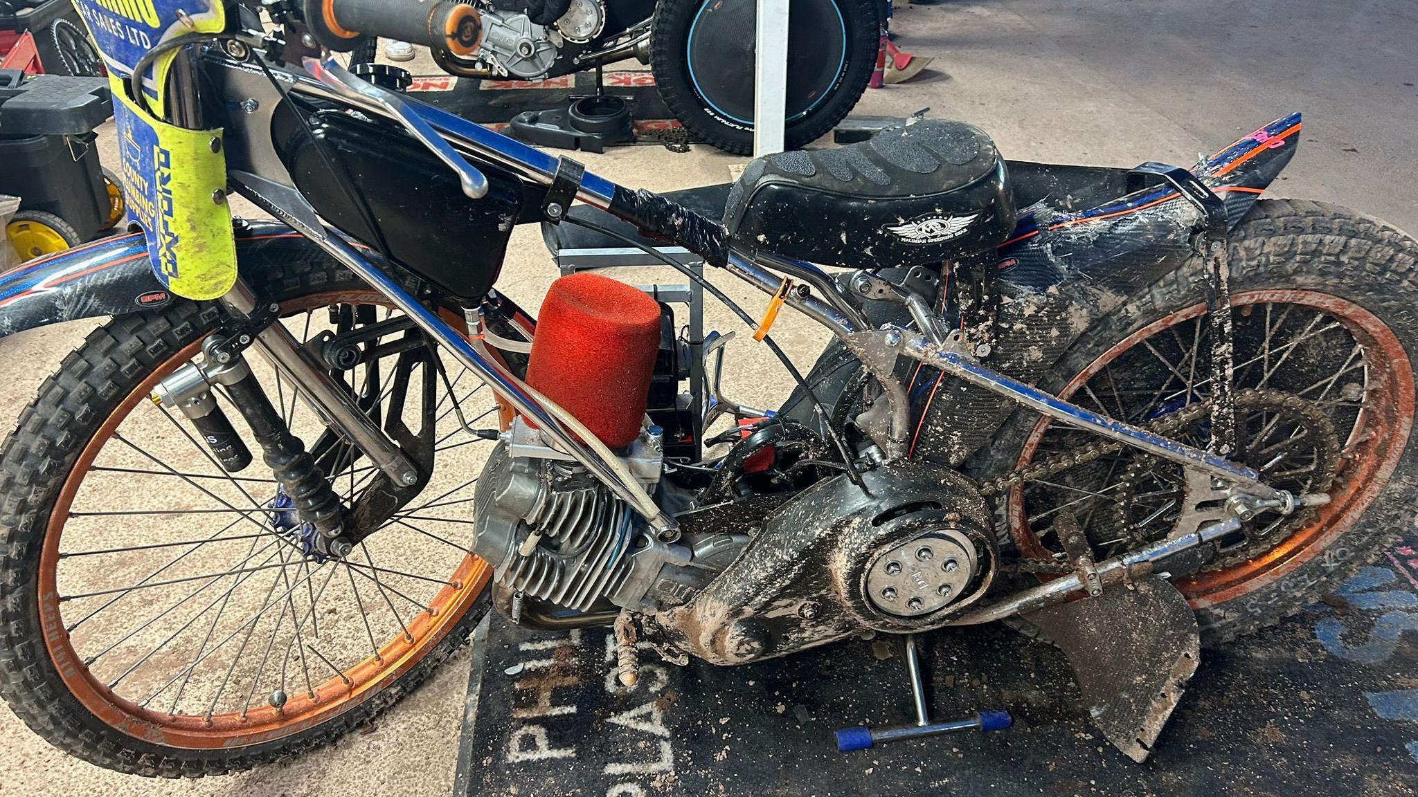 The damaged speedway bike propped up. It is covered in mud and dust.