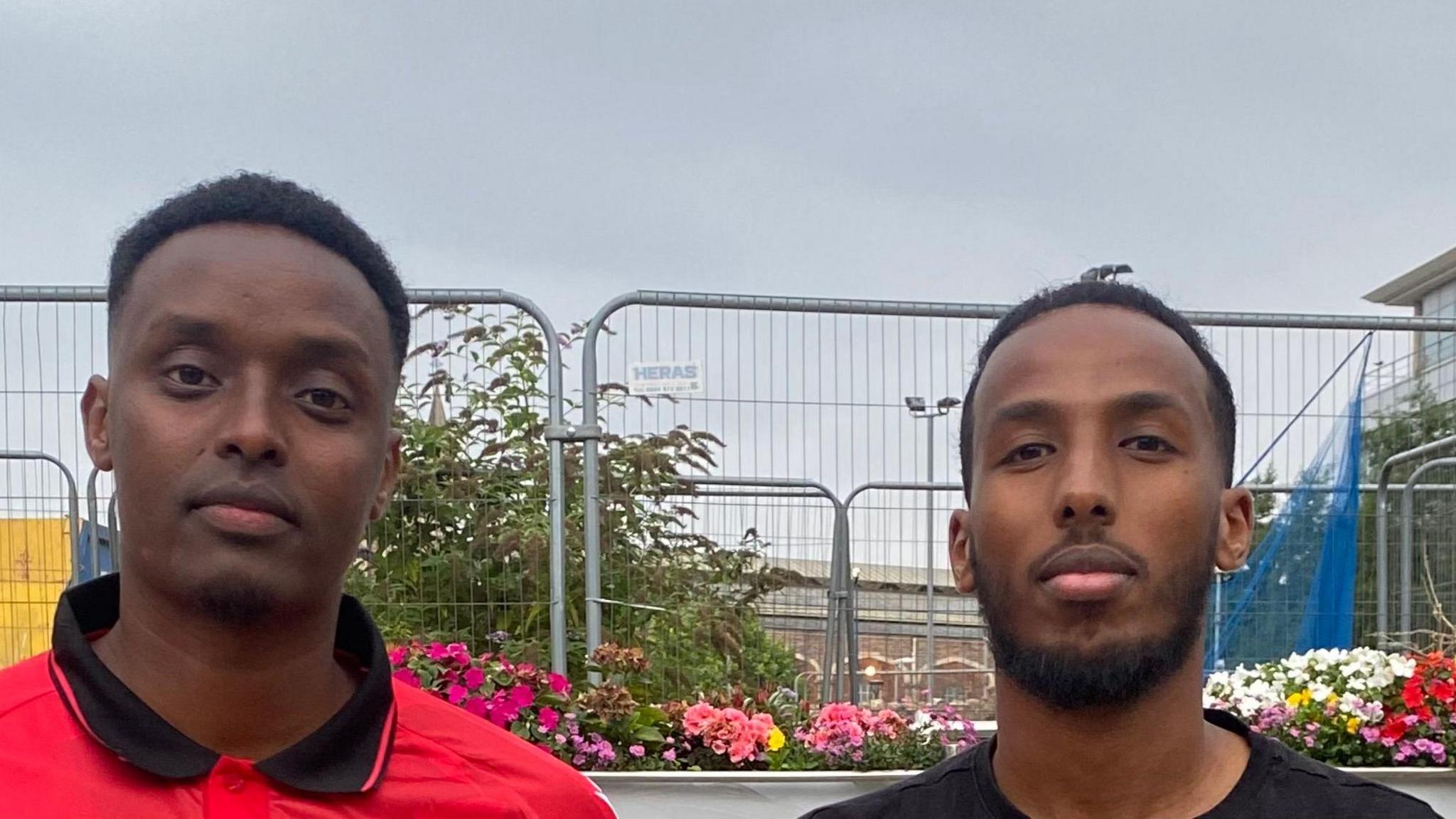 Two young men stand in front of flower beds and temporary fences. One wears a red polo shirt with a black collar, the other wears a black t-shirt.