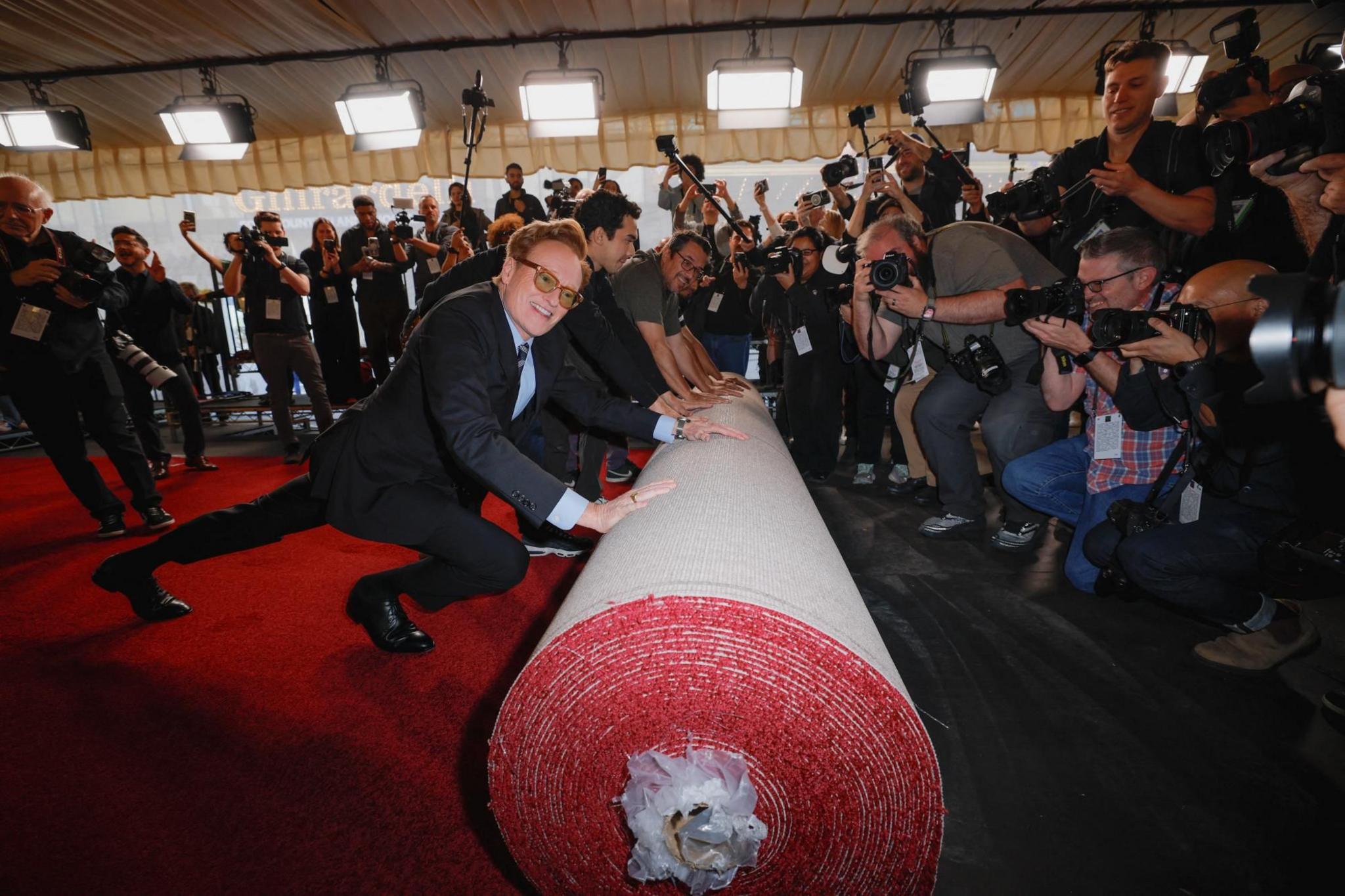 Conan O'Brien, wearing glasses and a suit, pushes a huge roll of red carpet out as a big crowd of photographers takes his picture