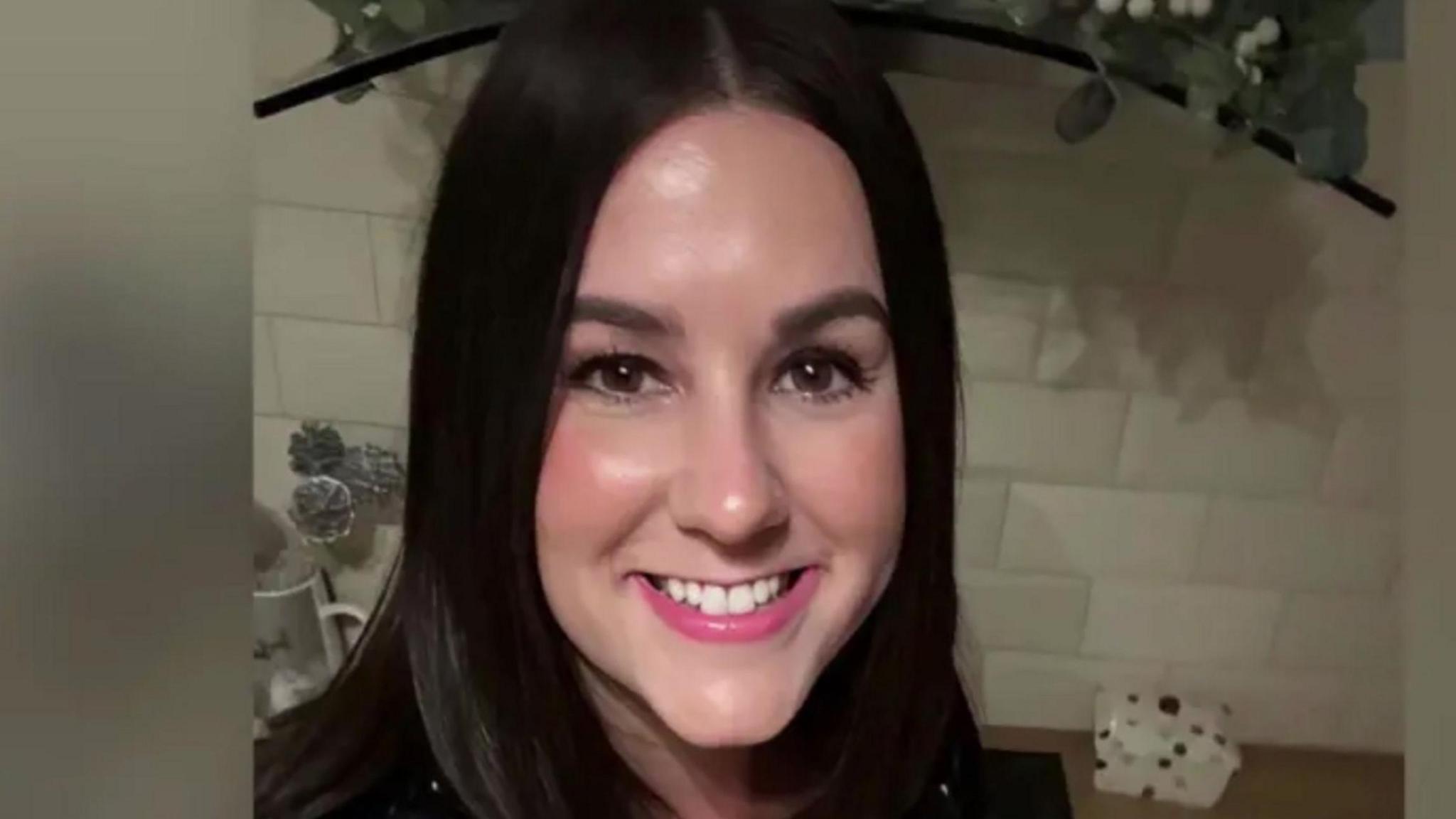 A smiling Lucy Connolly looking directly down the camera. She has shoulder length, dark brown hair and brown eyes. She is seen indoors, in what appears to be a kitchen, with a white tiled splashback and work surface behind her. The edge of a hob extractor fan can also be seen behind her.