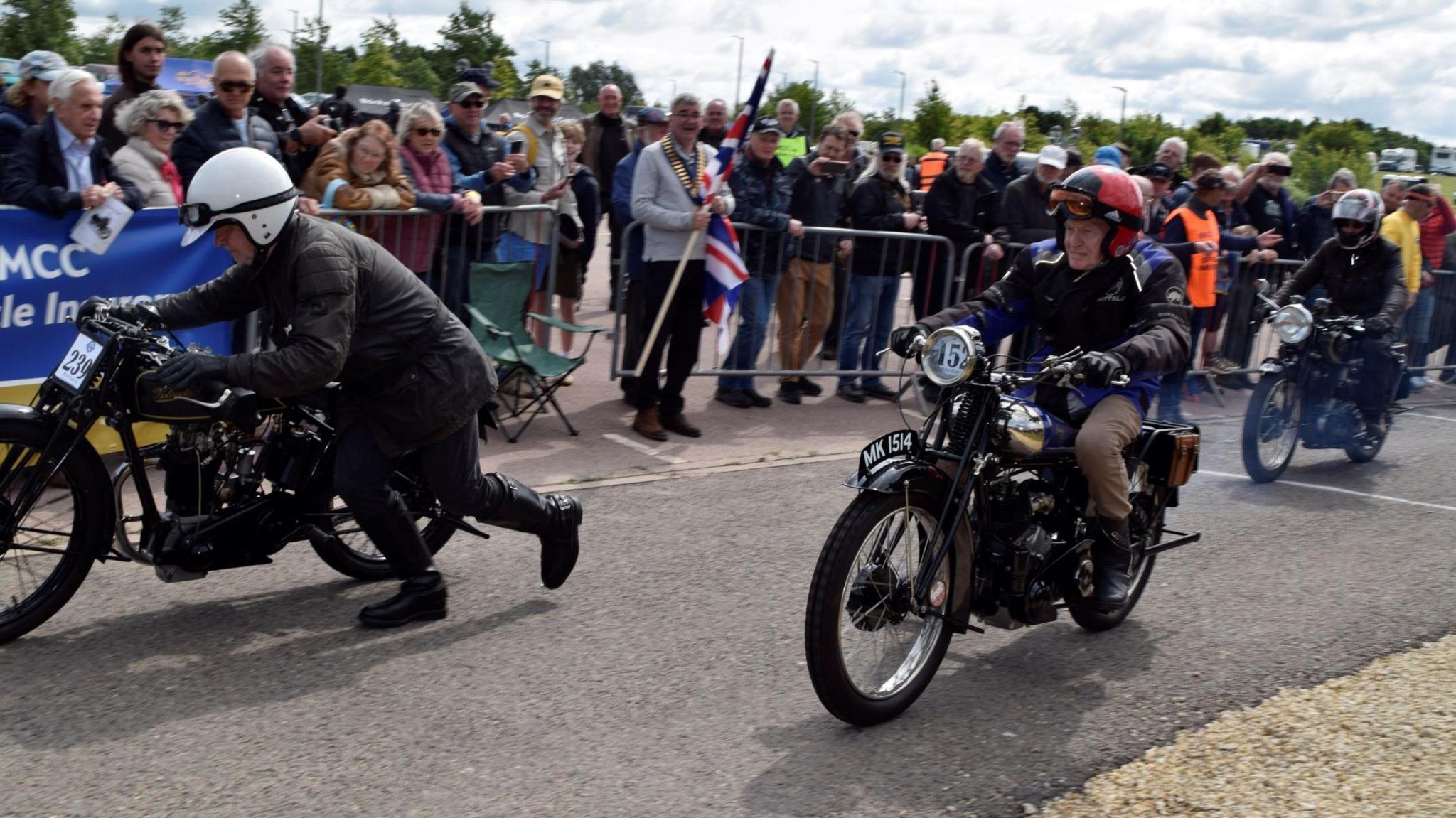  A man in a red cycling helmet is riding a vintage motorbike. Another motorcyclist is behind him and in front is another man who appears to be pushing his motorcycle. Crowds of people are watching from the other side of a barrier.