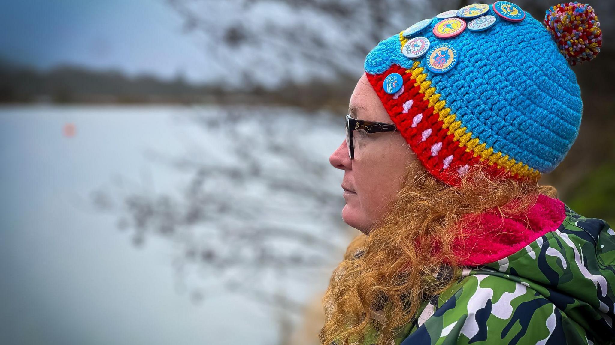 Sam Mitchell wearing a woollen hat and heavy coat with a hood after her swim. She is in profile looking out on the lake behind her