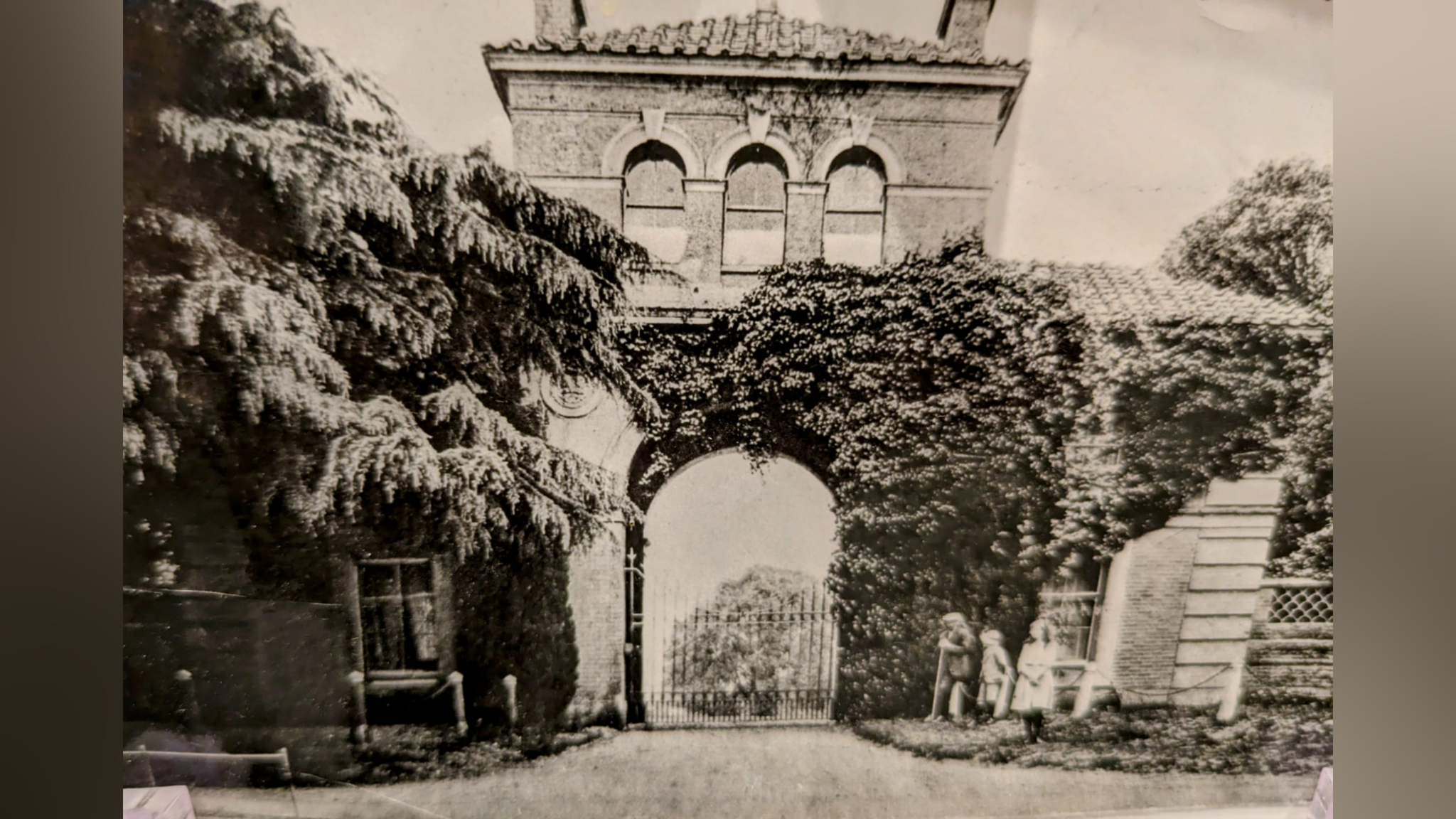 A black and white photo of a large archway which forms an entrance to Henham Park. Ivy covers part of the archway and three children stand in front of it. 