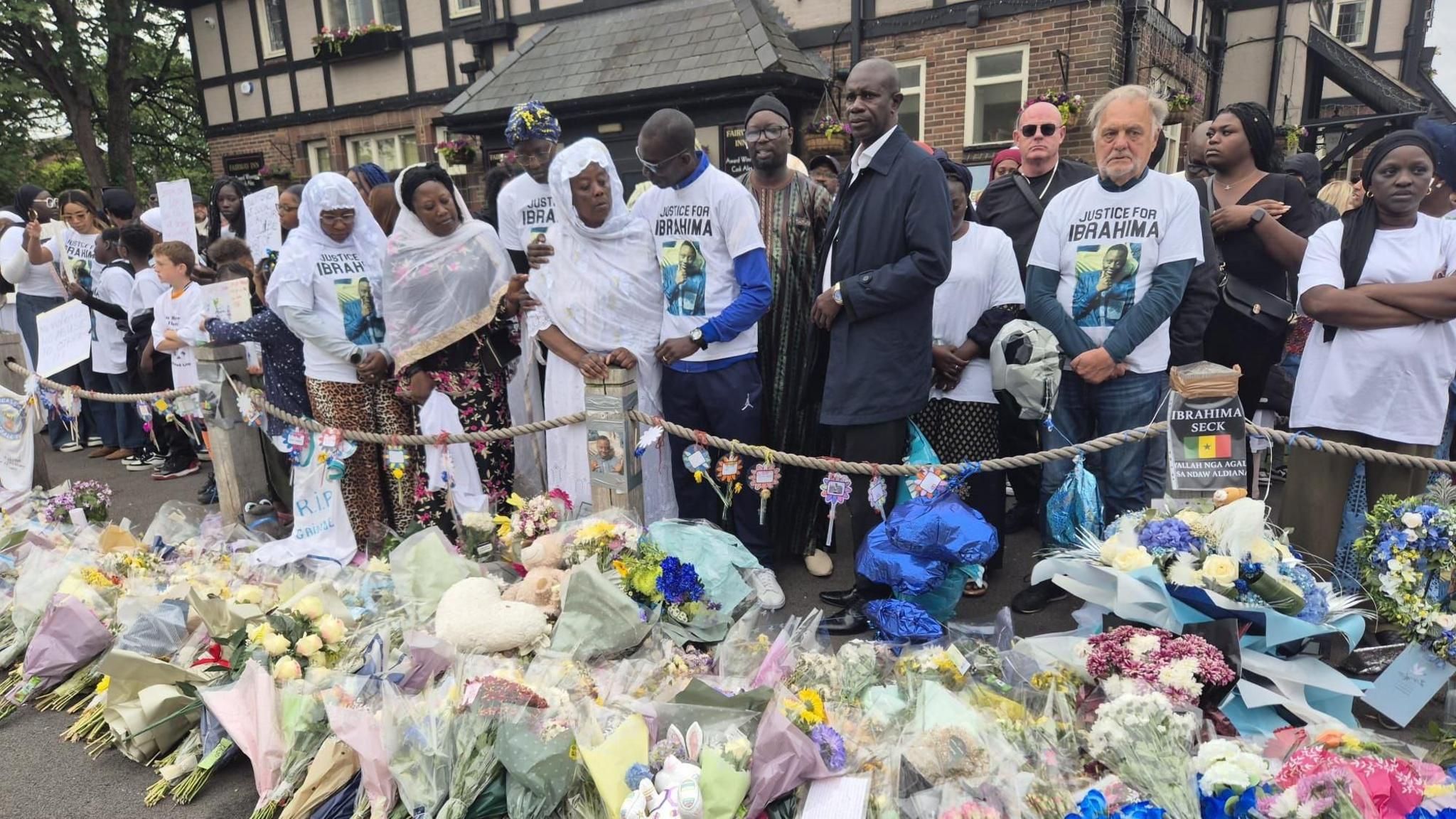 Ibrahima Seck's father clasps his arm around his wife while others stand beside them wearing white shirts with Ibrahima's picture. Floral tributes are laid out on the ground in front of them.