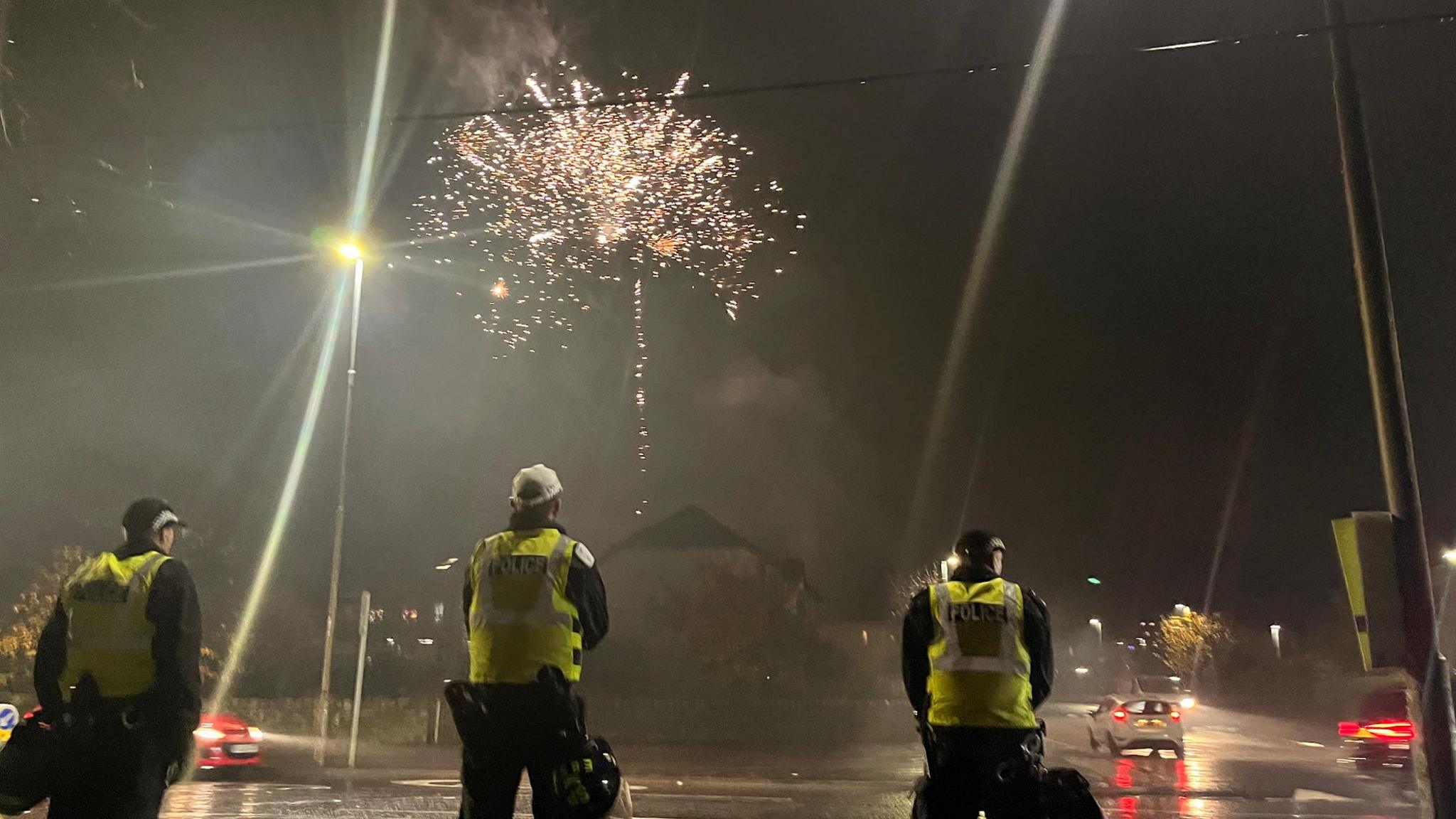 Police officer in high viz jackets on a wet street with fireworks lighting the sky behind them