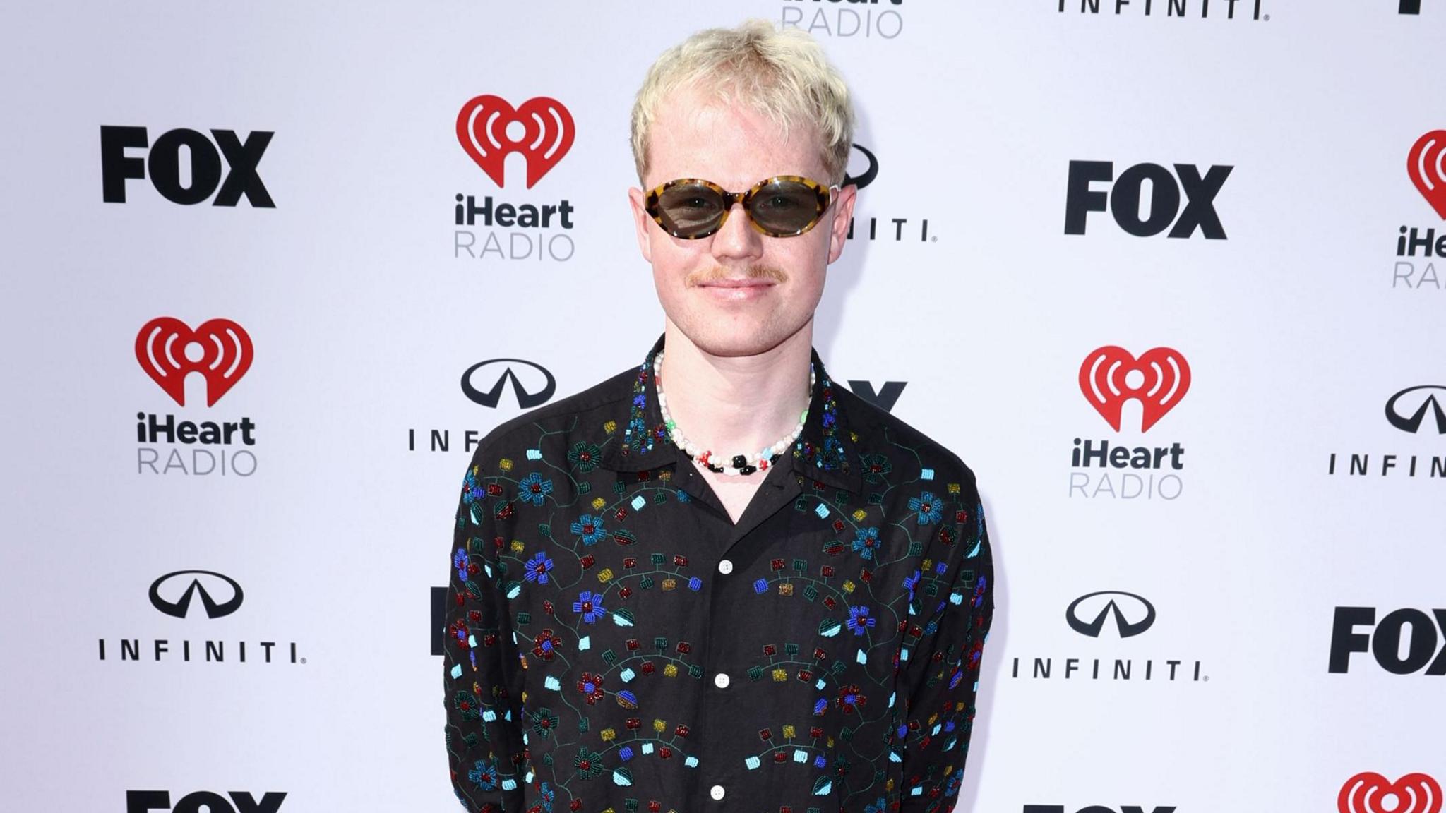 Lloyd Wakefield stands in front of a white backdrop that has Fox, iHeart Radio and Infiniti logos. He has short light blonde hair, a moustache and is wearing sunglasses. He is smiling at the camera and is wearing a black, floral beaded shirt with a colourful charm necklace underneath.