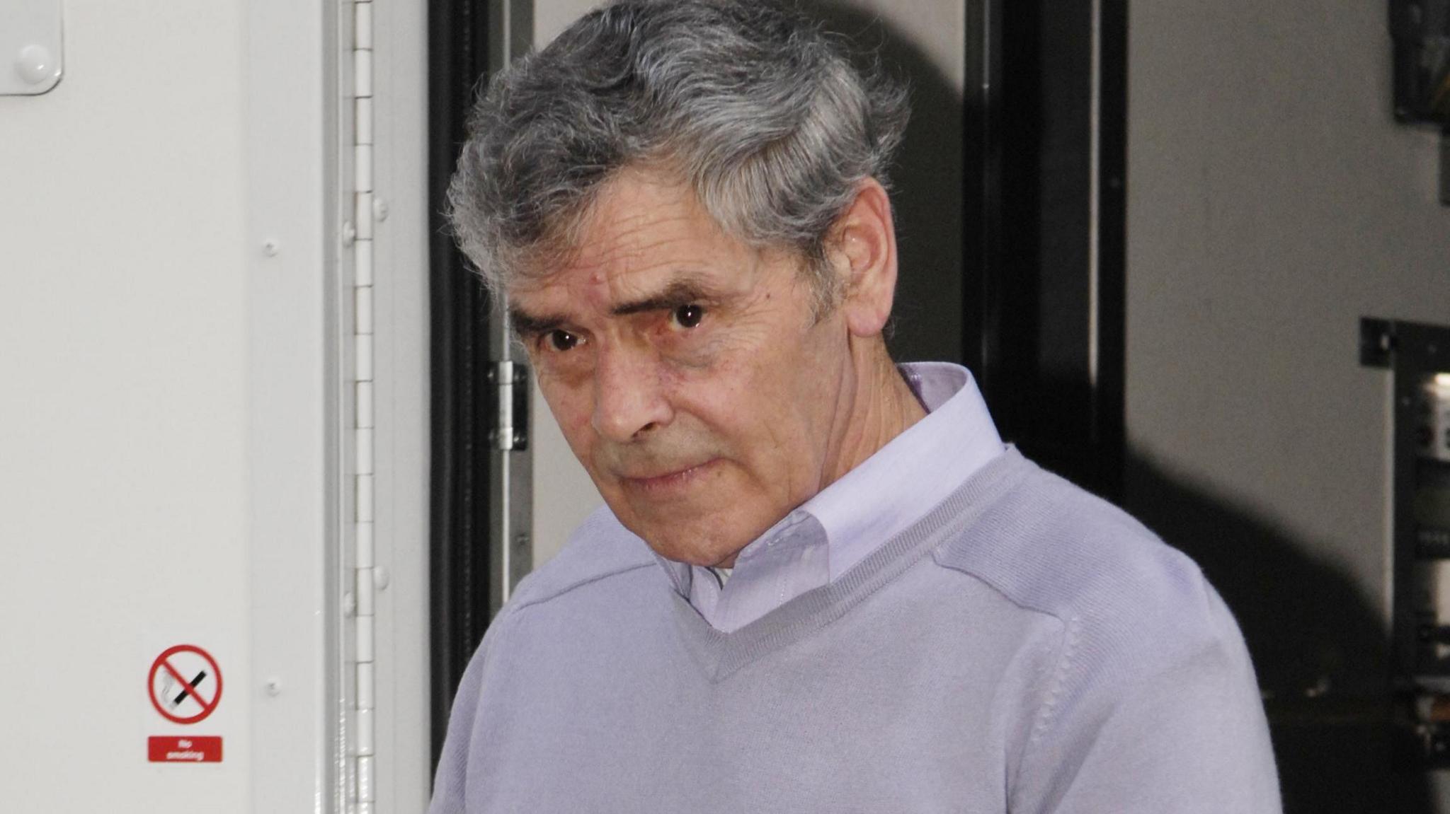 Peter Tobin comes out of a white van - he has grey hair and wears a lilac jumper over a lilac shirt. His expression is neutral.