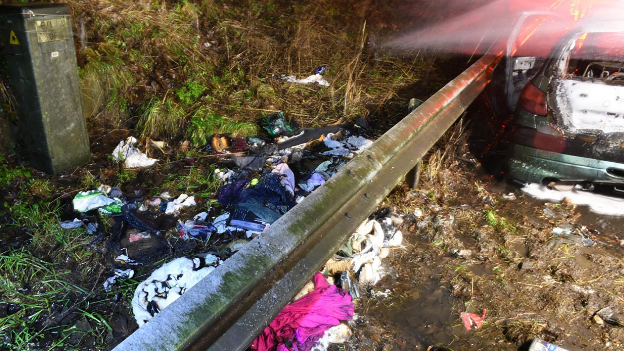 The twisted and charred remains of a car next to a metal crash barrier. There is a pile of semi-burned belongings strewn on a grass verge at the rear of the vehicle. 