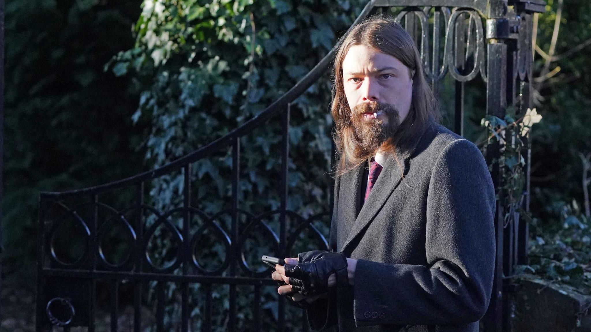 Benjamin Clarke outside Durham Crown Court. He has long dark hair and a beard, and is wearing a dark coat, black gloves and a dark red tie. He is looking into the camera and smoking a cigarette while holding a phone.