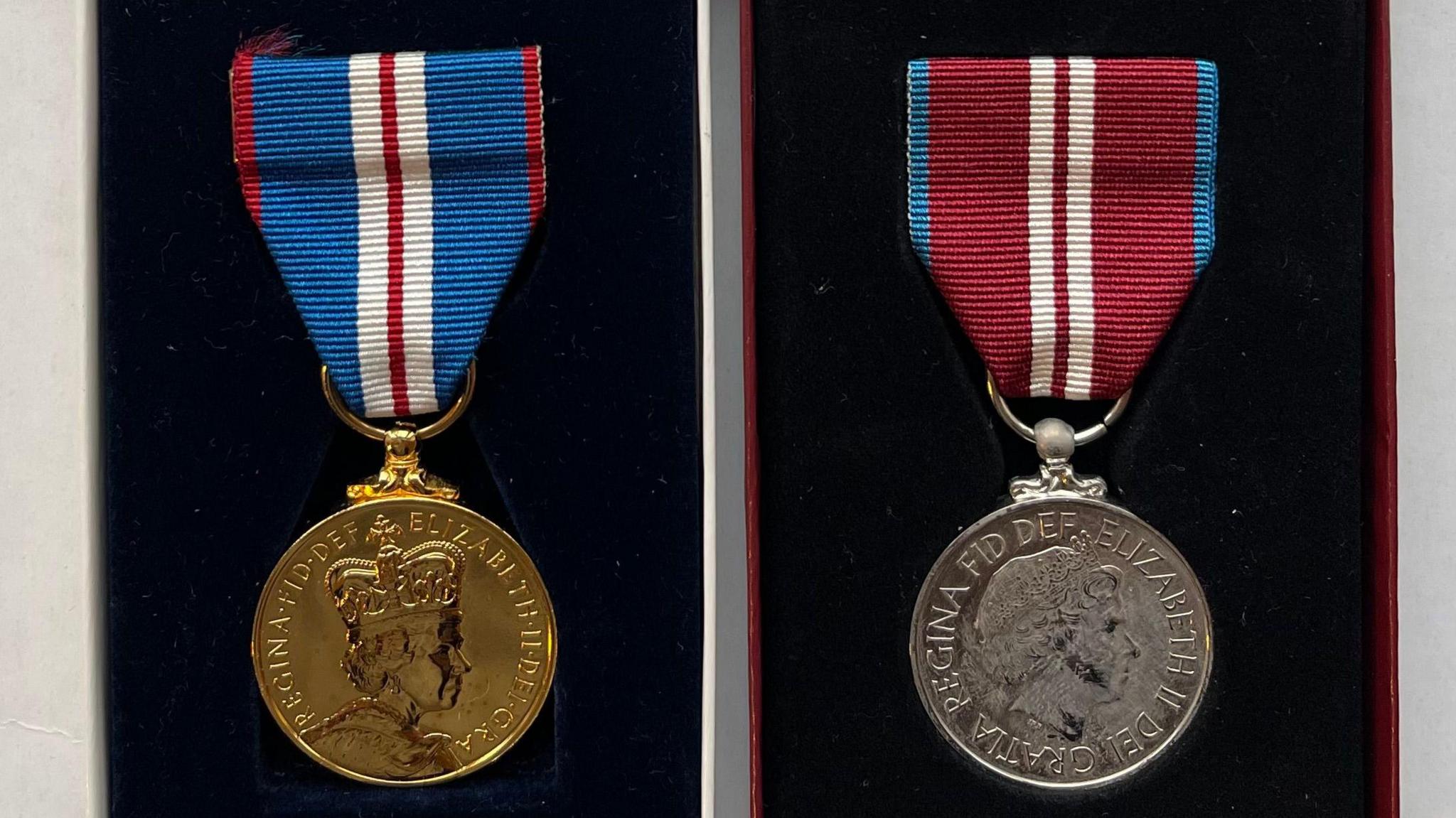 A gold medal featuring Queen Elizabeth's head and wording with blue, white and burgundy striped ribbon. Next to it is a matching medal in silver with a different striped ribbon but in the same colours. They are both on a black background.