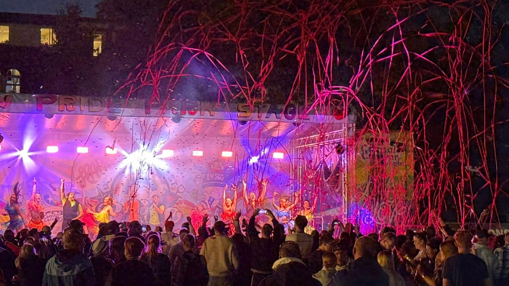 The stage is lit up at night in bright purple and red colours.  Red streamers have fired into the air and performers have their hands up in the air as if they are at the end of the show.