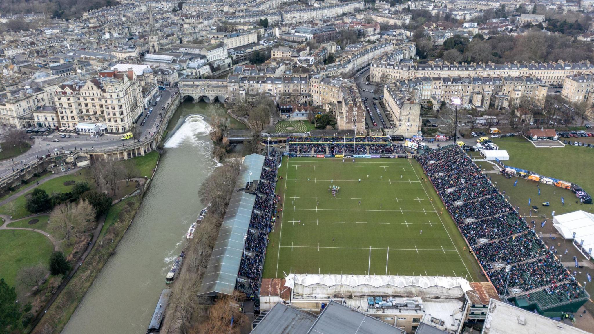 An aerial shot of the Recreation Ground, home of Bath Rugby. The River Avon is visible running alongside the stadium, as is much of Bath's historic city centre. There is a match going on and the stands in the stadium are full of spectators