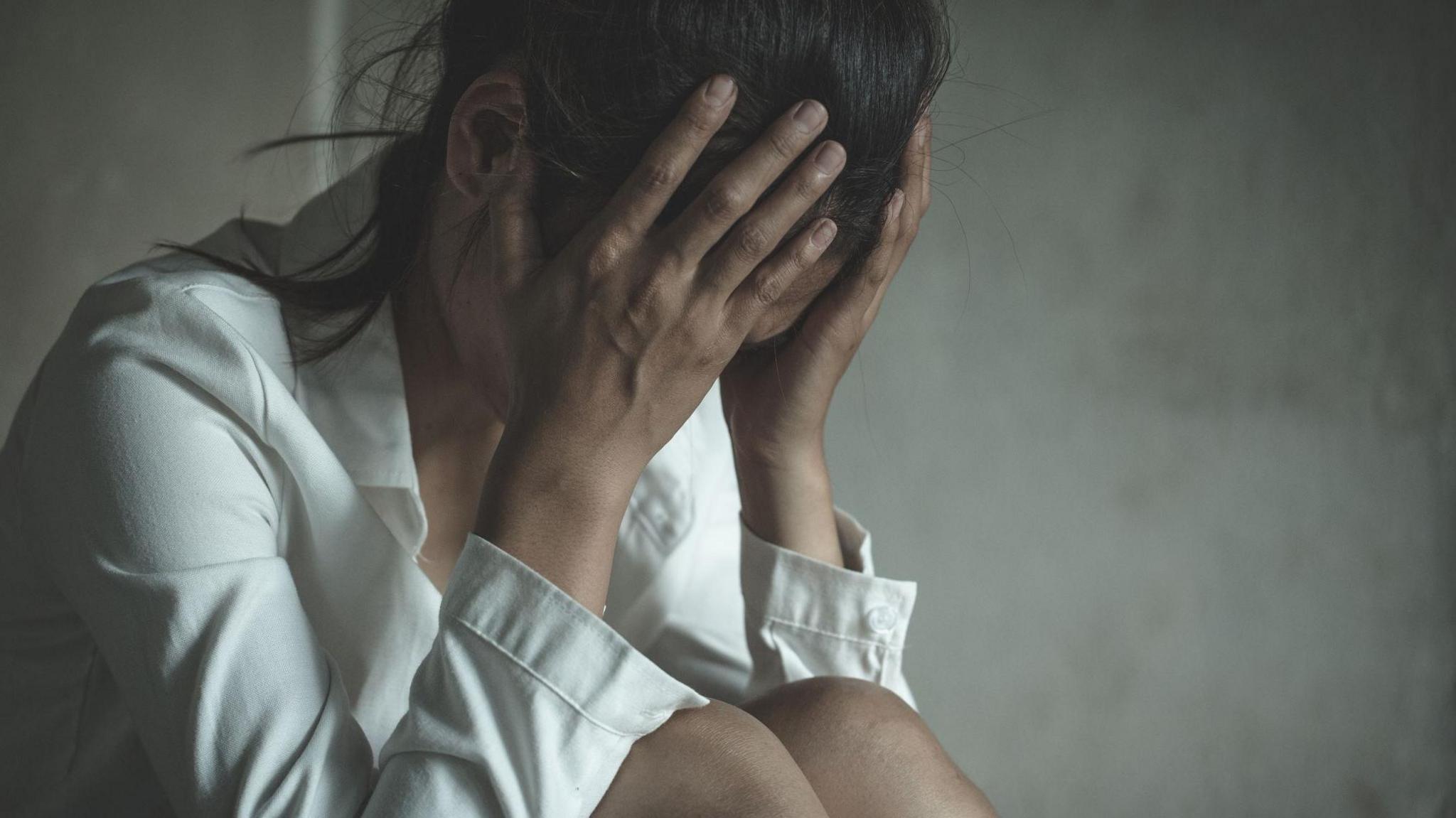 A woman with her head in her hands. She has dark hair which is tied back and she is wearing a white shirt.