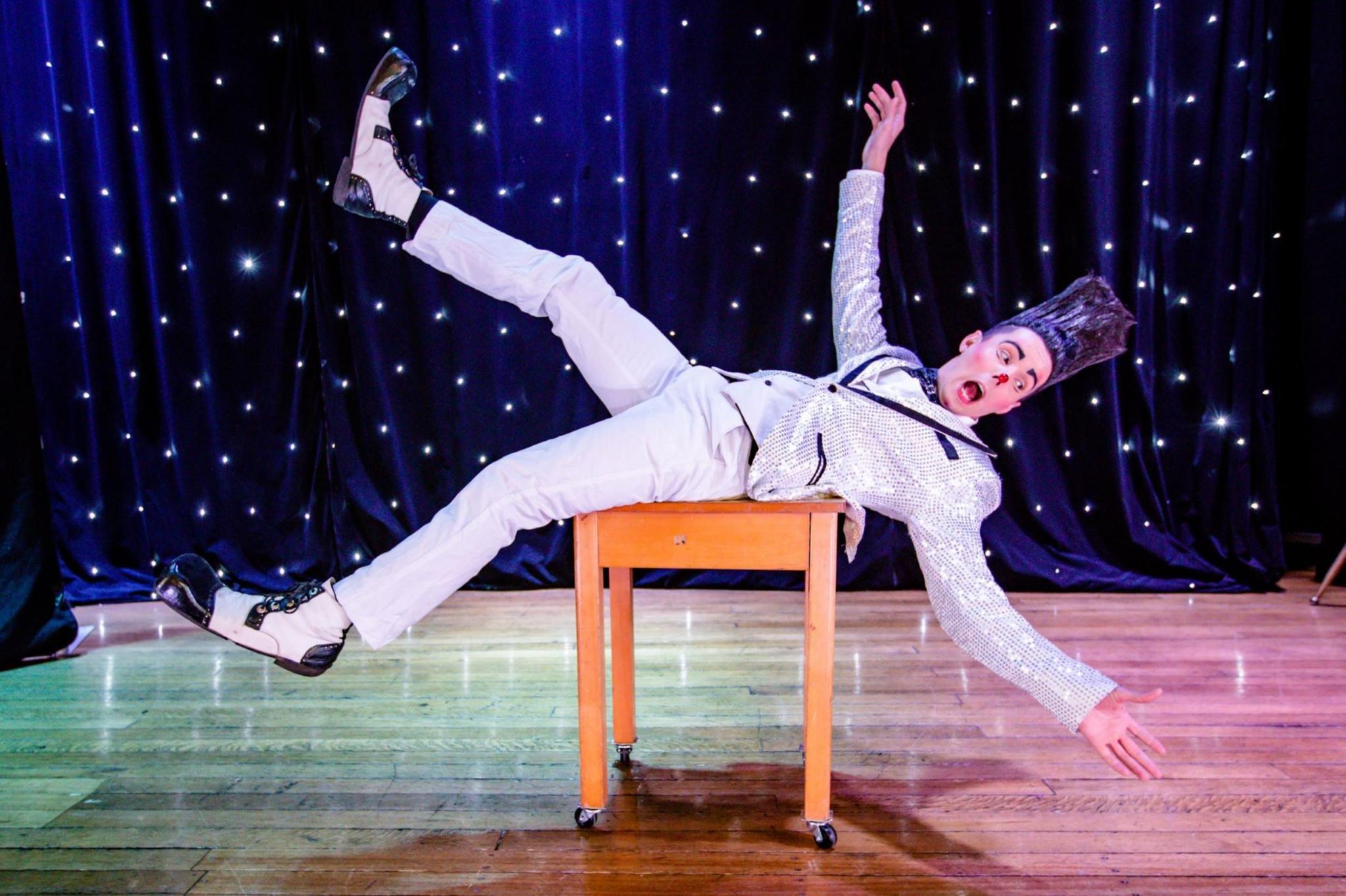 A performer with very high gelled hair wears a silver suit and lies with his arms sprawled out on a desk. He is on a stage with a dark blue backdrop with twinkling lights on it.