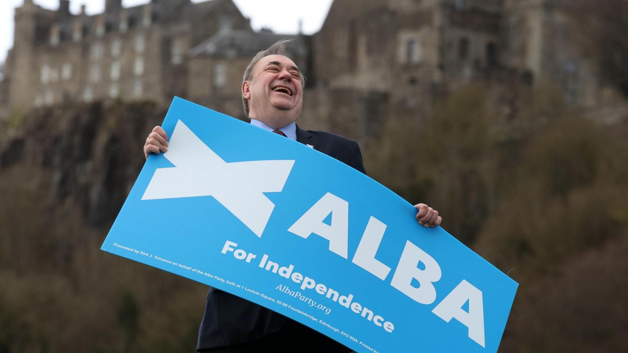 Alex Salmond laughing and holding a blue Alba party poster, with Edinburgh Castle in the background