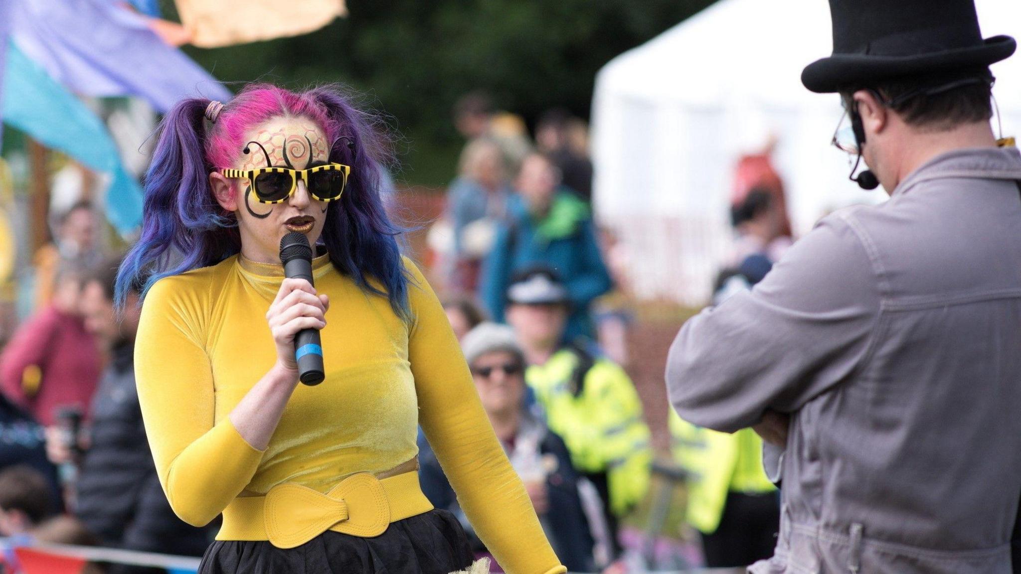 A woman with two hair bunches that are dyed pink and purple is singing into a microphone. She is wearing black and yellow stripy sunglasses and has black and orange honeycomb style face paint. She is wearing a yellow top and belt