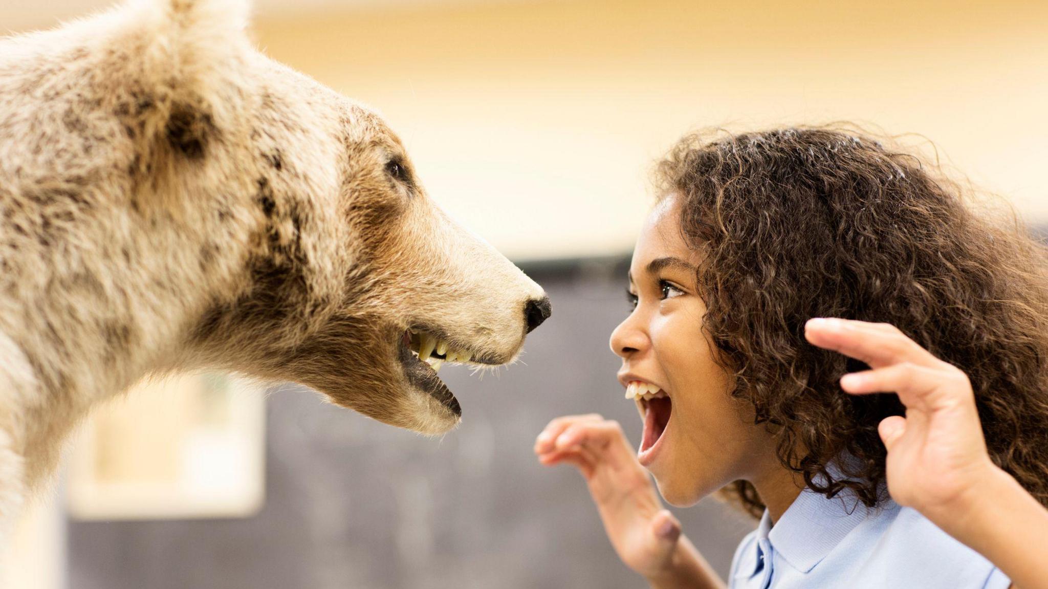 A girl with curly brown hair pretending to roar at a stuffed bear in a museum.