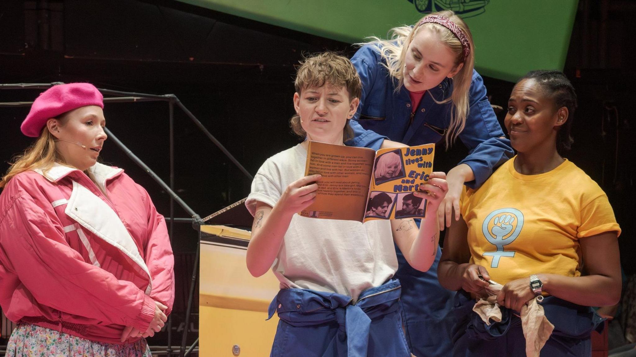 Four performers stand together on stage in a theatrical scene. One wears a pink beret, pink jacket, and floral skirt; another holds a booklet titled 'Jenny lives with Eric and Martin' and is dressed in blue overalls. A third person, also in overalls, looks over their shoulder, while the fourth wears a yellow t-shirt with a feminist symbol and holds cloth or paper. Industrial props and signage are visible in the background.