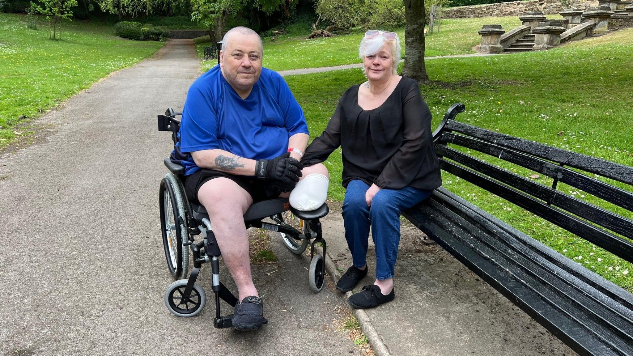 A man is sitting in a wheelchair in a park holding his wife's hand who is sitting on a bench next to him. His left leg has been amputated below the knee. He is wearing shorts and a blue polo shirt and his wife is wearing jeans and a black top.