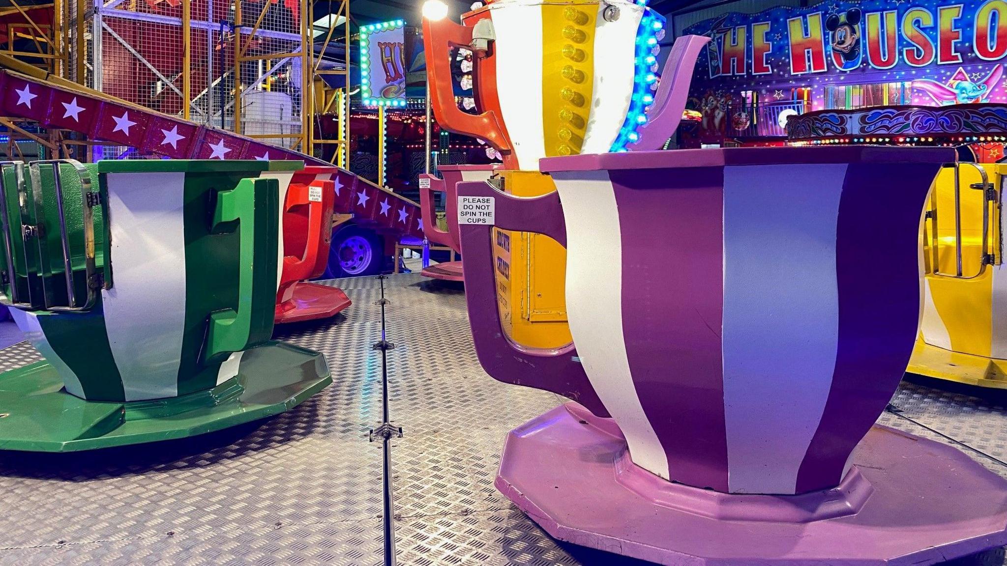 Giant teacups that rotate are stationary in front of a House of Fun. The teacups are green and white striped, purple and white striped and red and white striped. The helter skelter is in the background.