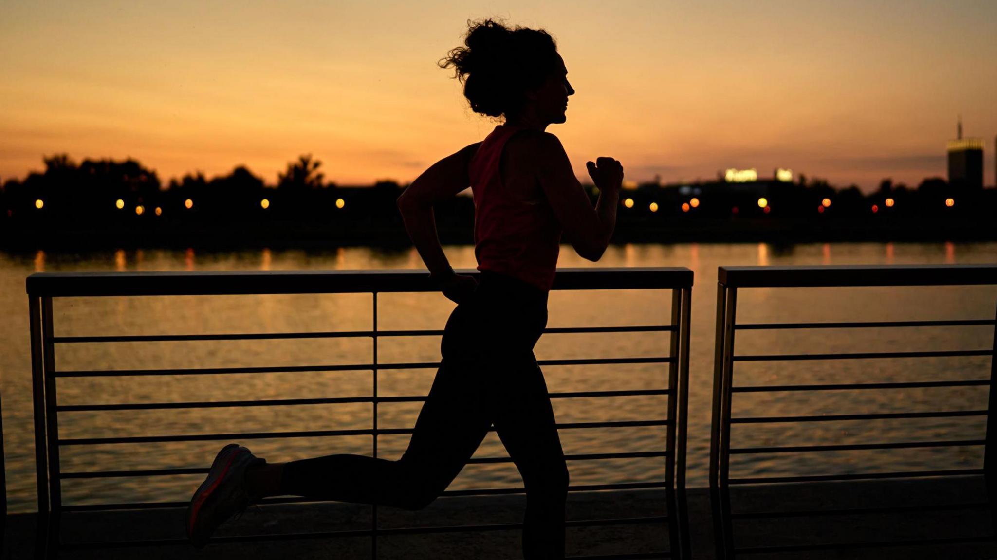 The image shows a person jogging along a waterfront promenade during sunset. The silhouette of the runner is clearly visible against the warm orange and pink hues of the sky. In the background, there is a calm body of water reflecting the sunset colours, with a row of trees and distant buildings illuminated by small lights. The foreground features a metal railing that runs parallel to the water, adding structure to the scene.