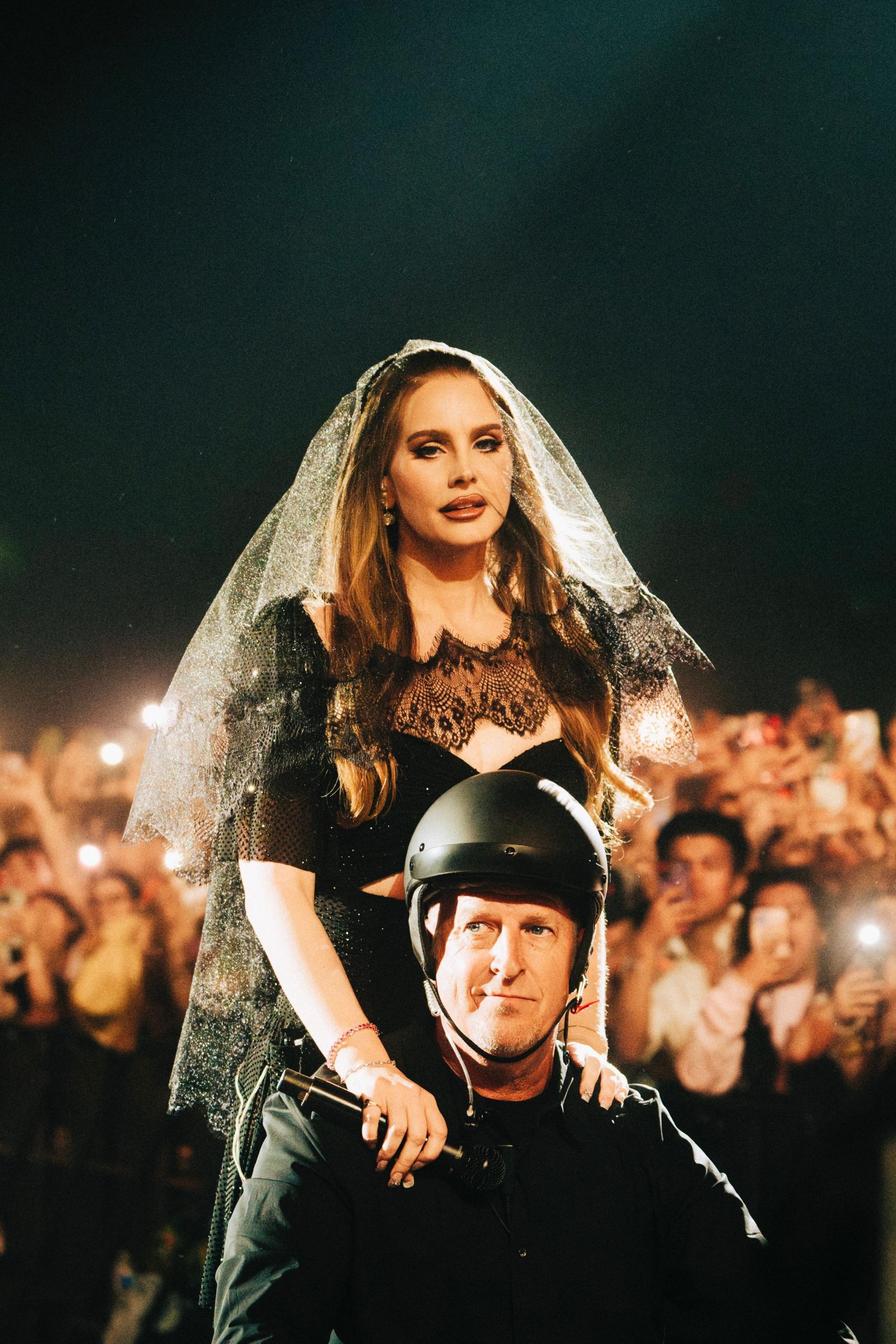 Lana Del Ray in what looks like black bridal gear sits on the shoulders of a man in a black helmet and jacket as crowds photograph and film them on mobile phones.