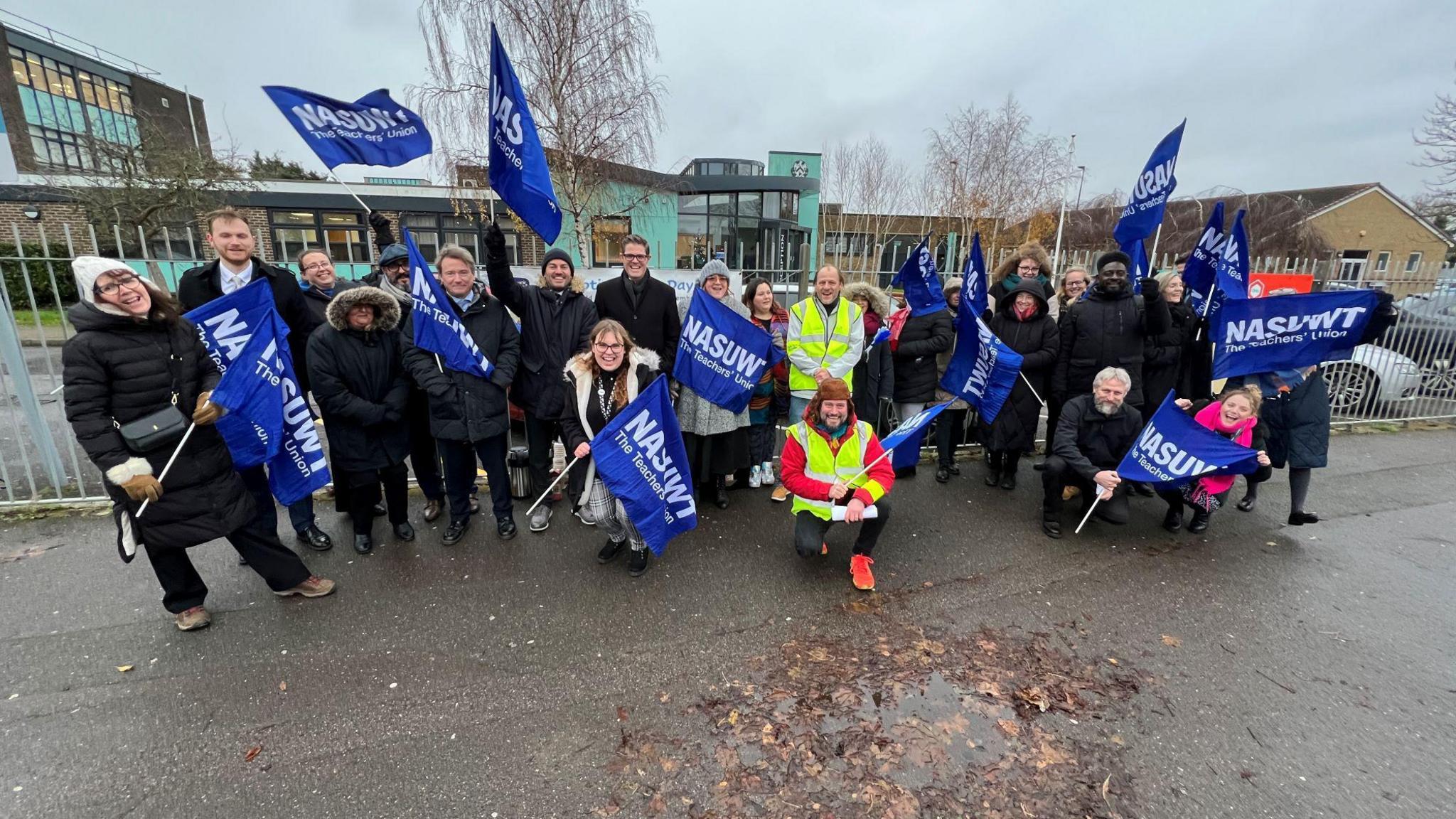 Group of people in winter clothing holding union flags outside a school fence.