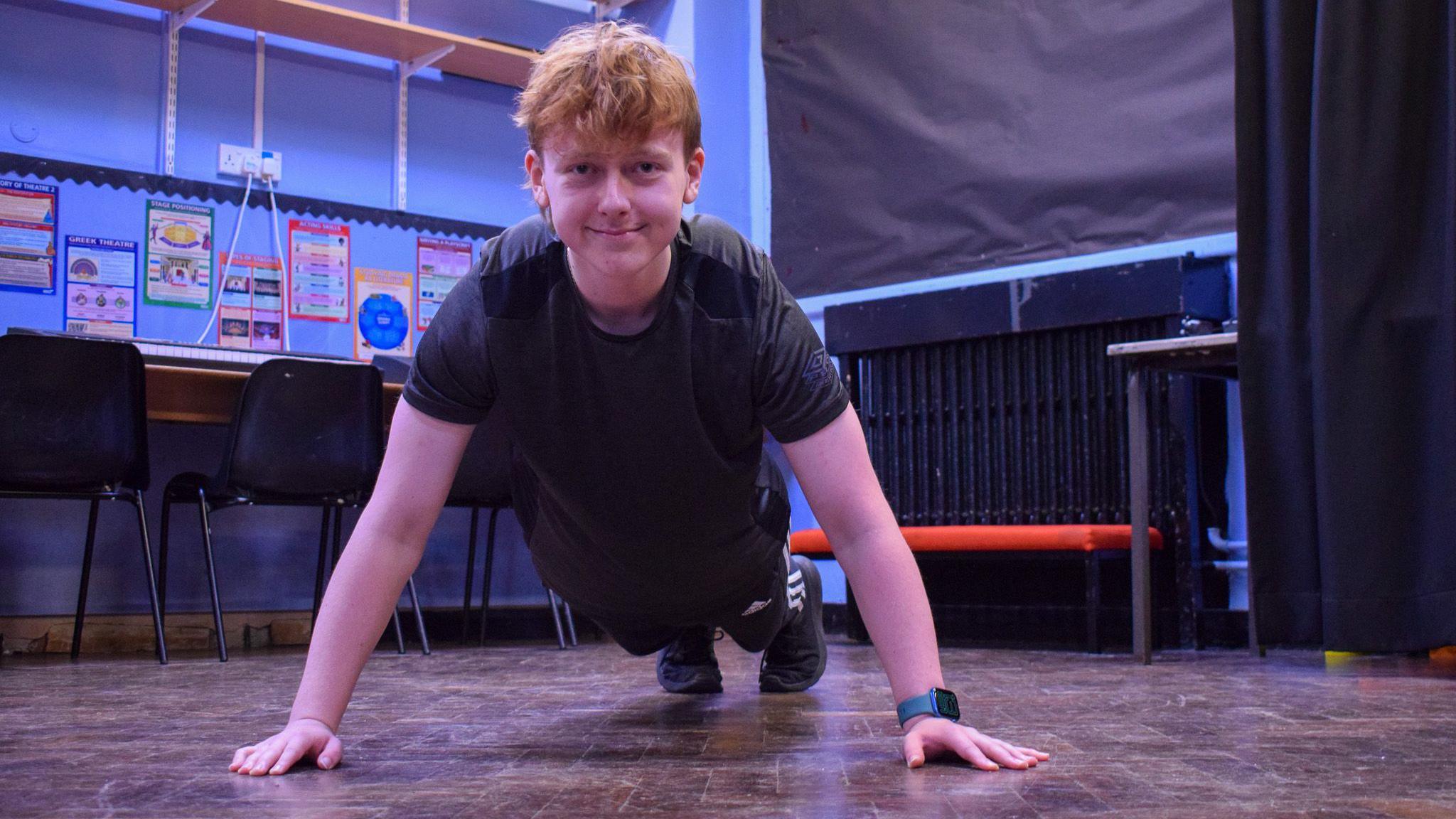 Adam is lying in the push-up position and is smiling at the camera, he is wearing a black t-shirt and black joggers with trainers. 