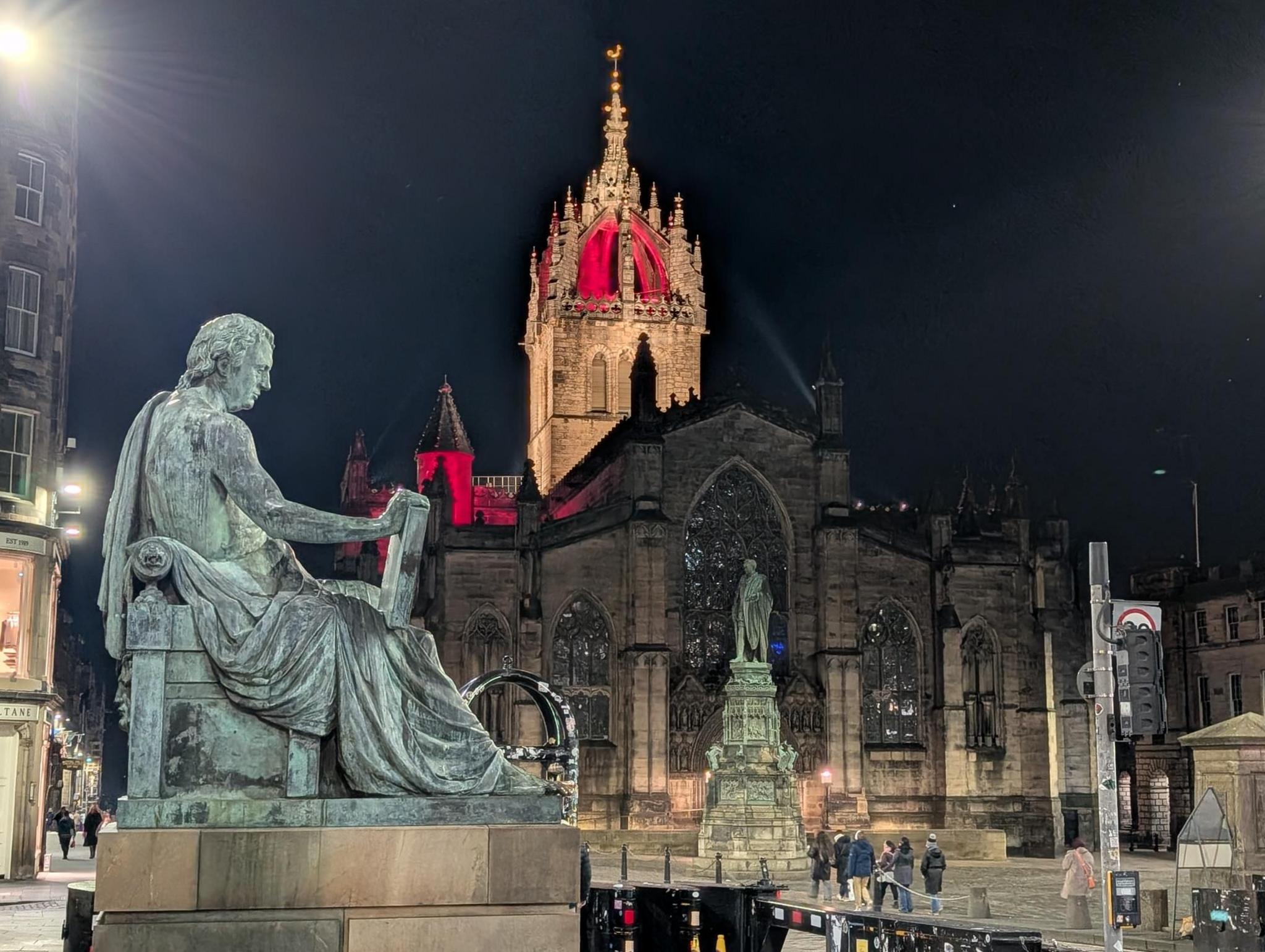 A grand Gothic-style cathedral dominates the scene, its ornate spire dramatically lit in red against the night sky. In the foreground, a bronze statue sits on a stone pedestal, overlooking the bustling street below. The area is illuminated by bright city lights, with people walking near the historic building.