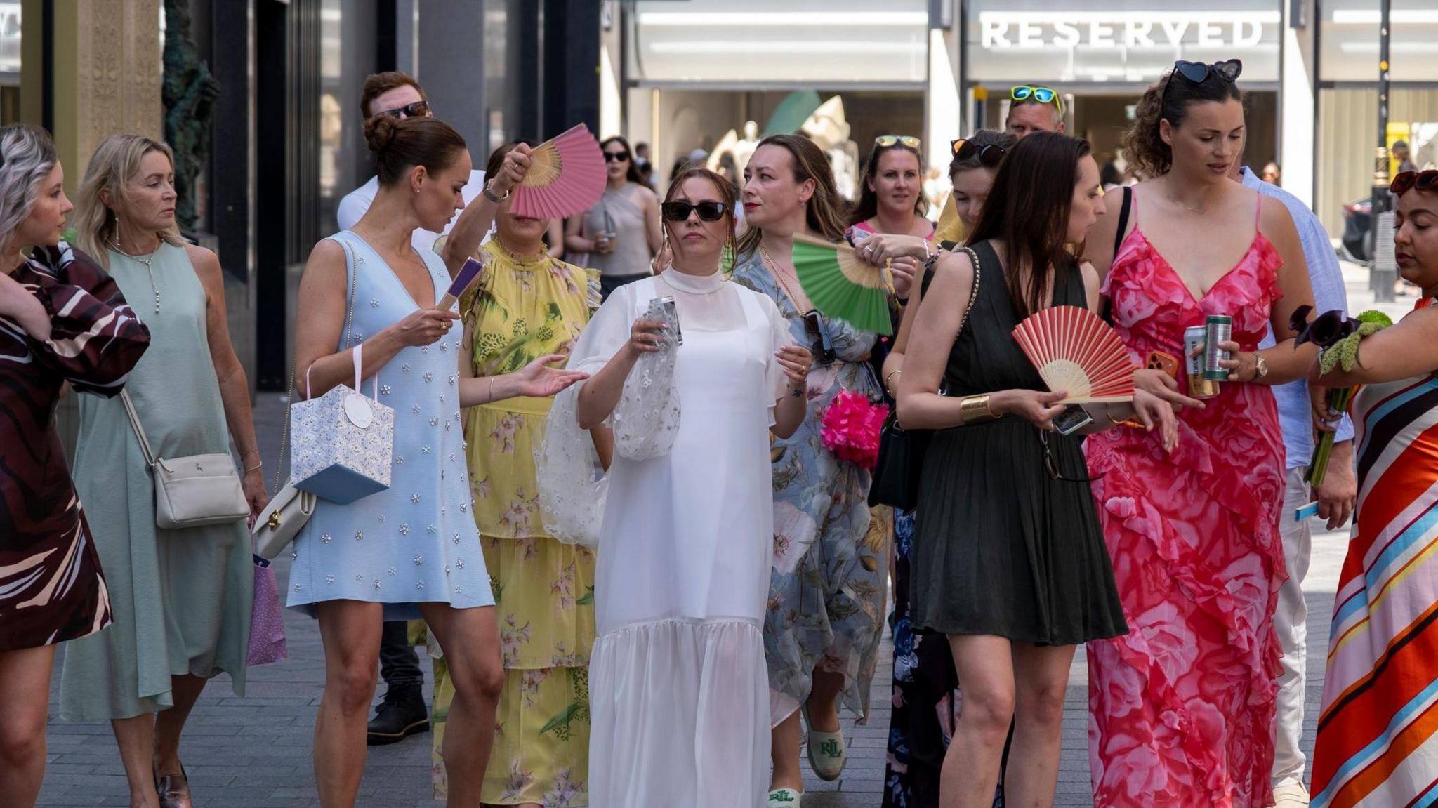 A bride walks through the centre of London with female guests in brightly coloured dresses surround her trying to fan her to keep cool
