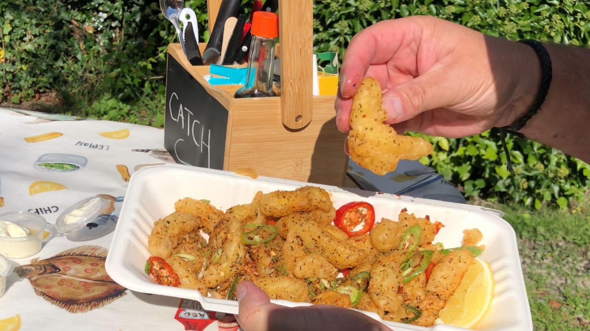A man's hand can be seen holding a chip with one hand and a container of chips with the other. The chips have herbs and cut up pieces of fresh chilli on them.