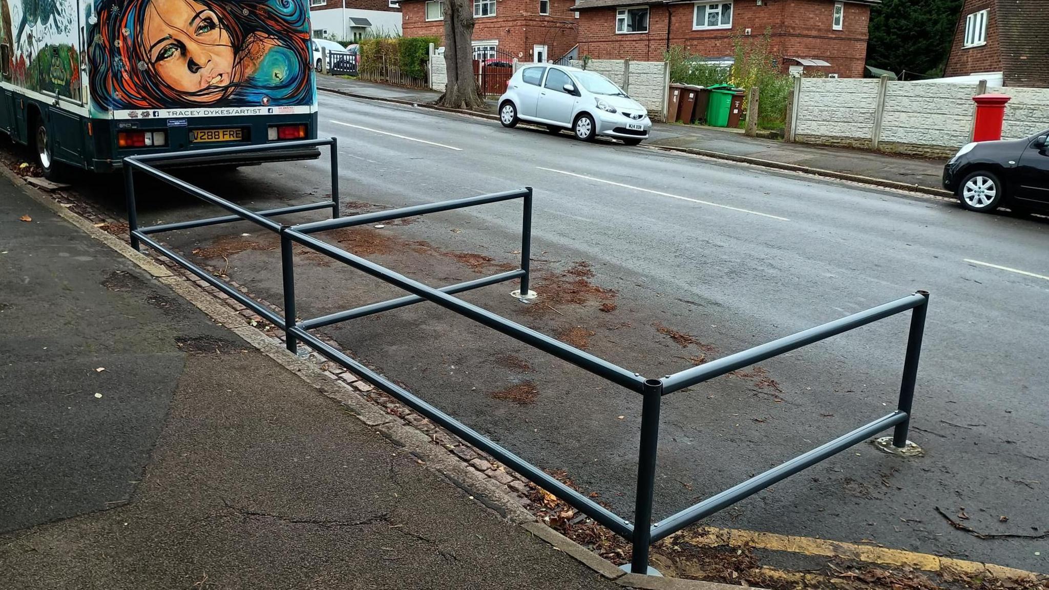 Grey tubular metal fencing, divided into two bays, sited next to a pavement and opening onto the roadway