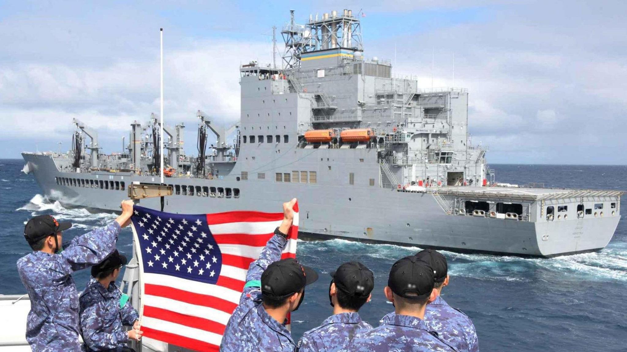 Crewmembers wave an American flag from another boat on the ocean as the US Navy oiler USNS Harvey Milk
