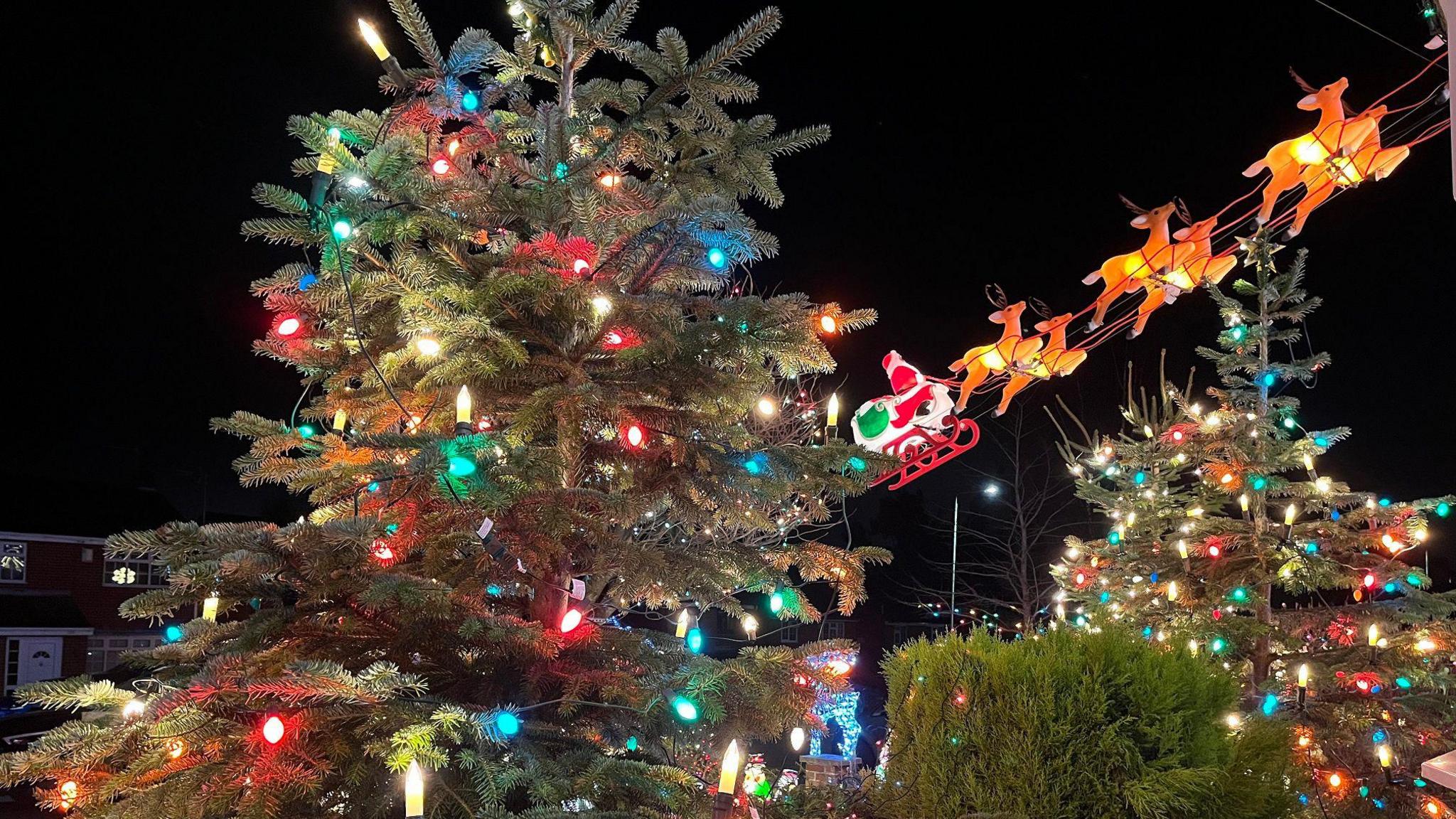 two christmas trees outside a house with warm glowing coloured light bulbs, plus Santa and his reindeer in the background flying over the house 